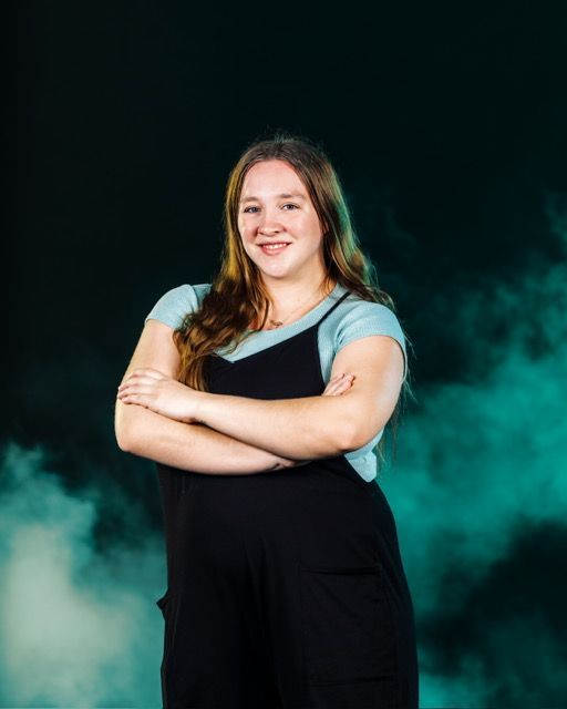 Woman in black Adidas shirt and pants, hands on hips, smiles in front of stage lights.