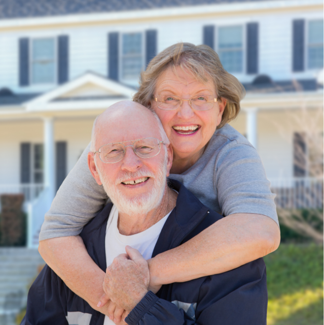 Smiling couple embraces in front of a white house. Woman has arms around man's neck.