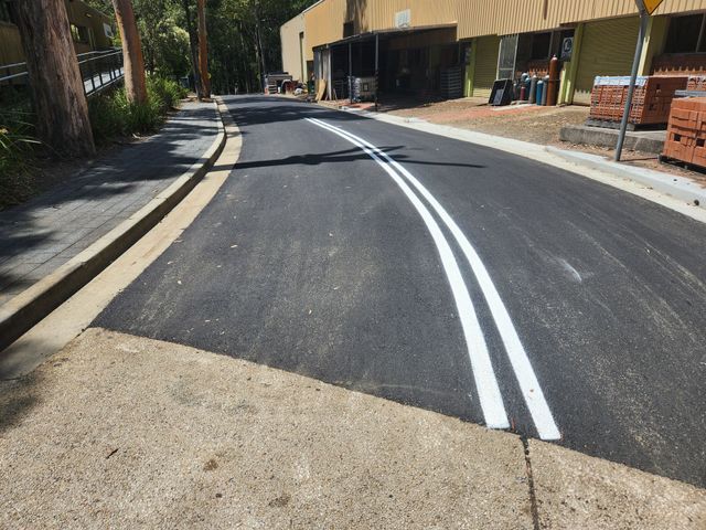 A newly paved asphalt road featuring fresh white double lines, bordered by a concrete curb and a building on the right.