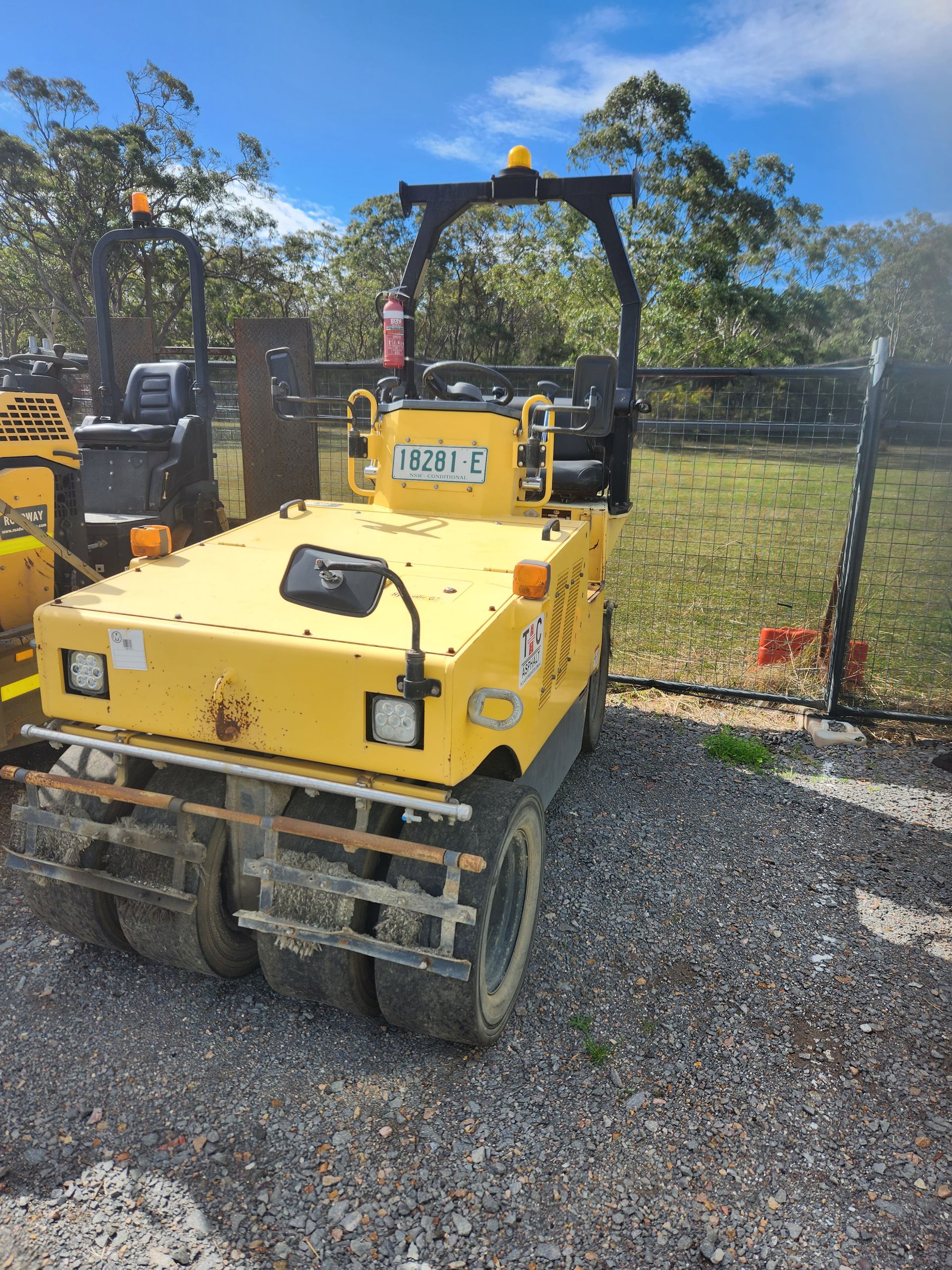 A yellow pneumatic tire roller parked on gravel under a blue sky, with a partial view of another machine on the left.