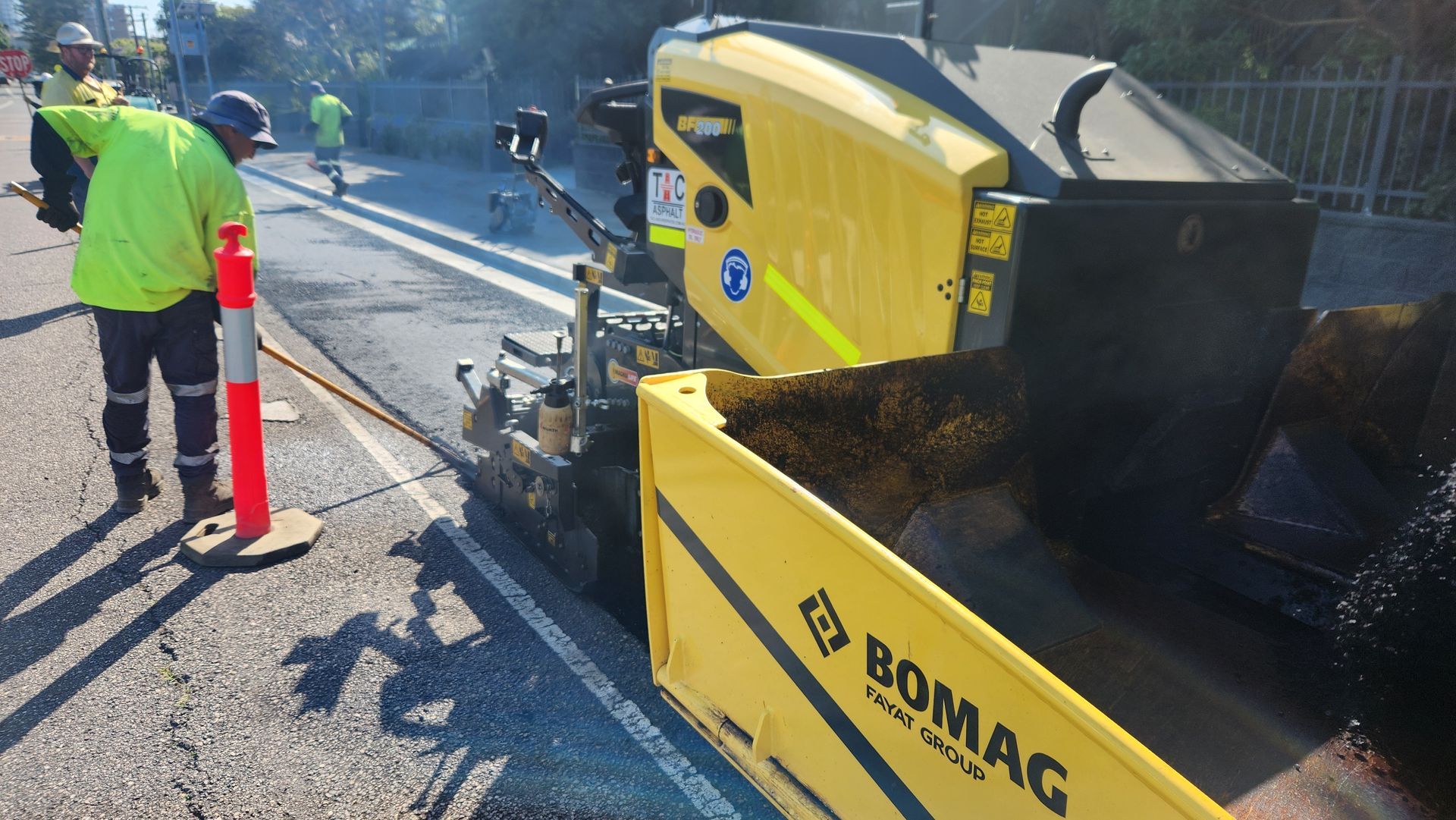 A worker in high-visibility gear operates a yellow BOMAG asphalt paver on a road construction site.