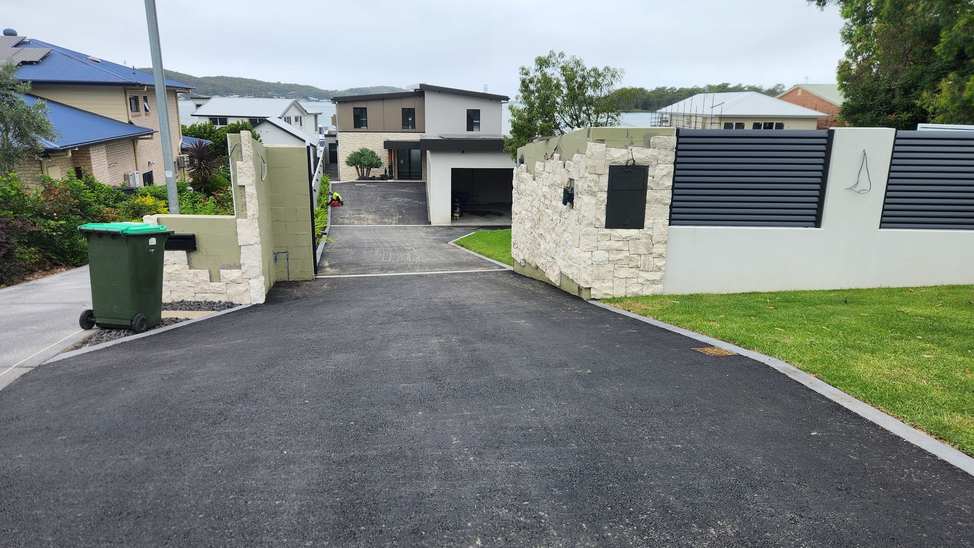 A paved driveway leading to a modern two-story house with a white stone wall and dark metal fencing at the entrance.