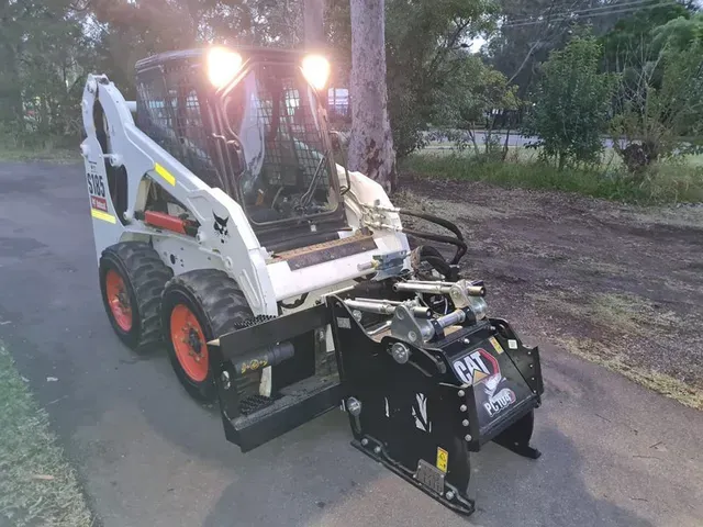 A white Bobcat skid steer loader with glowing headlights and an attached black CAT stump grinder on a paved driveway.