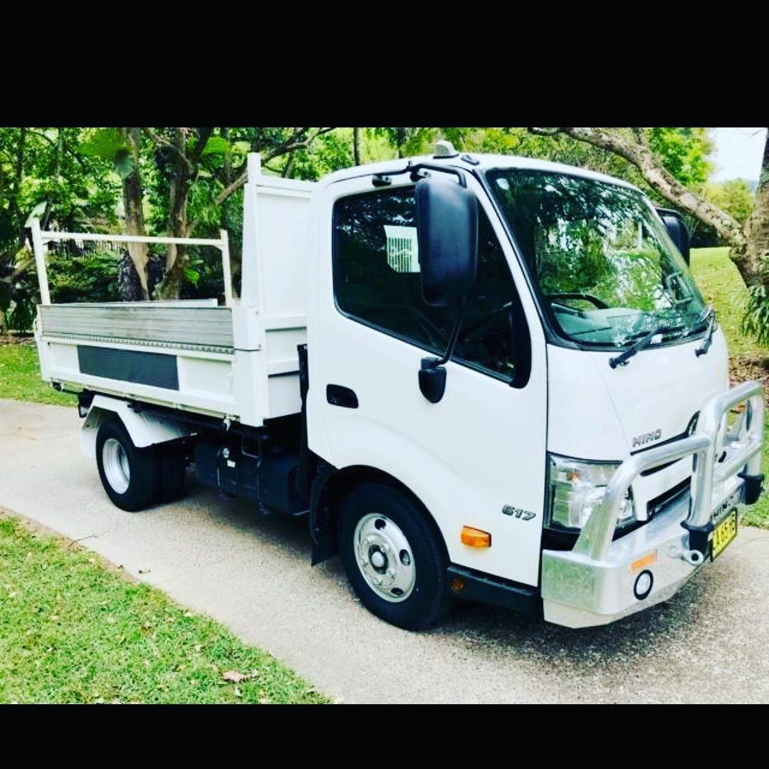 A white Hino light-duty tipper truck with a metal bull bar parked on a paved driveway surrounded by trees.