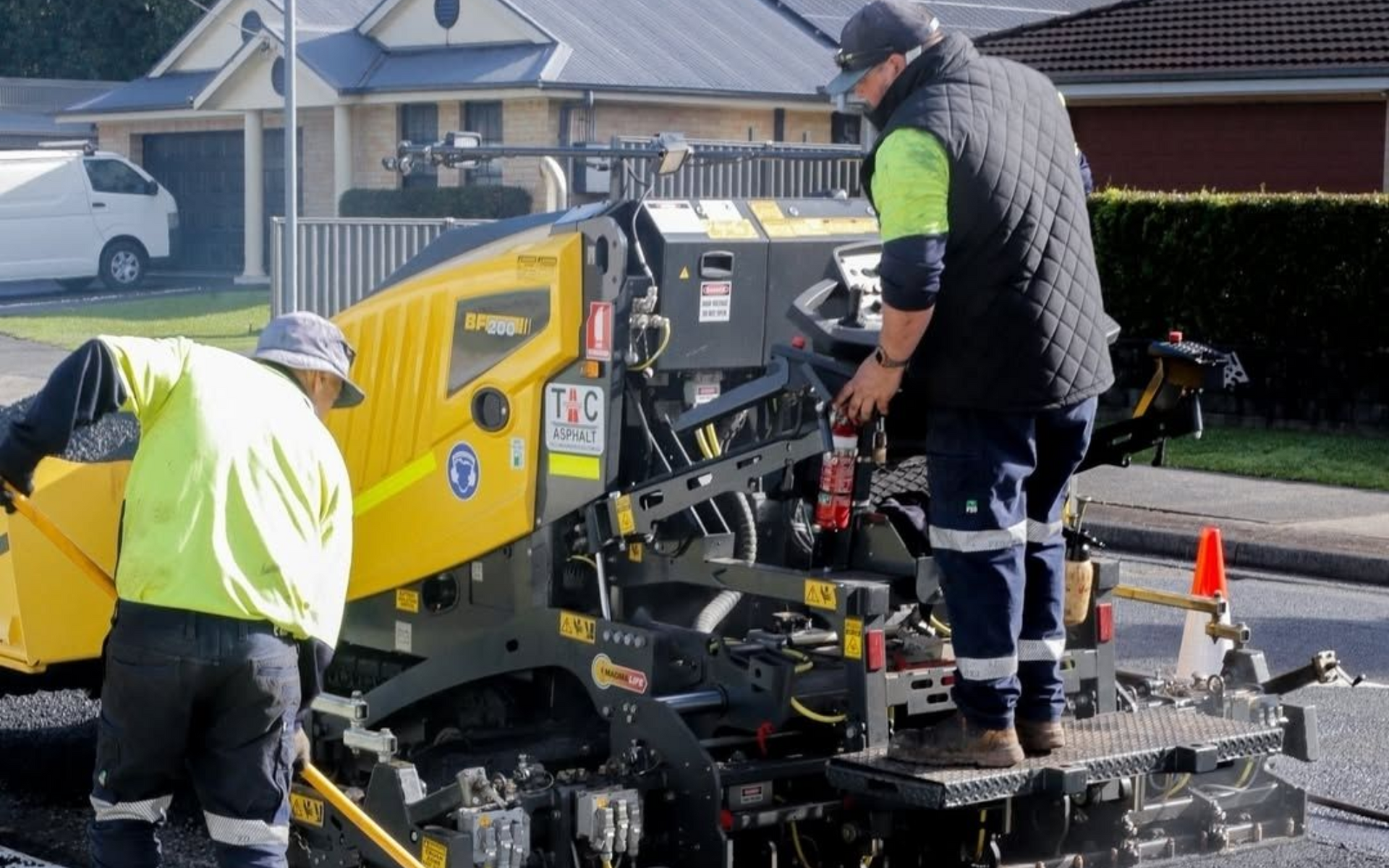 Two construction workers in high-visibility gear operate a yellow paving machine on a suburban street.