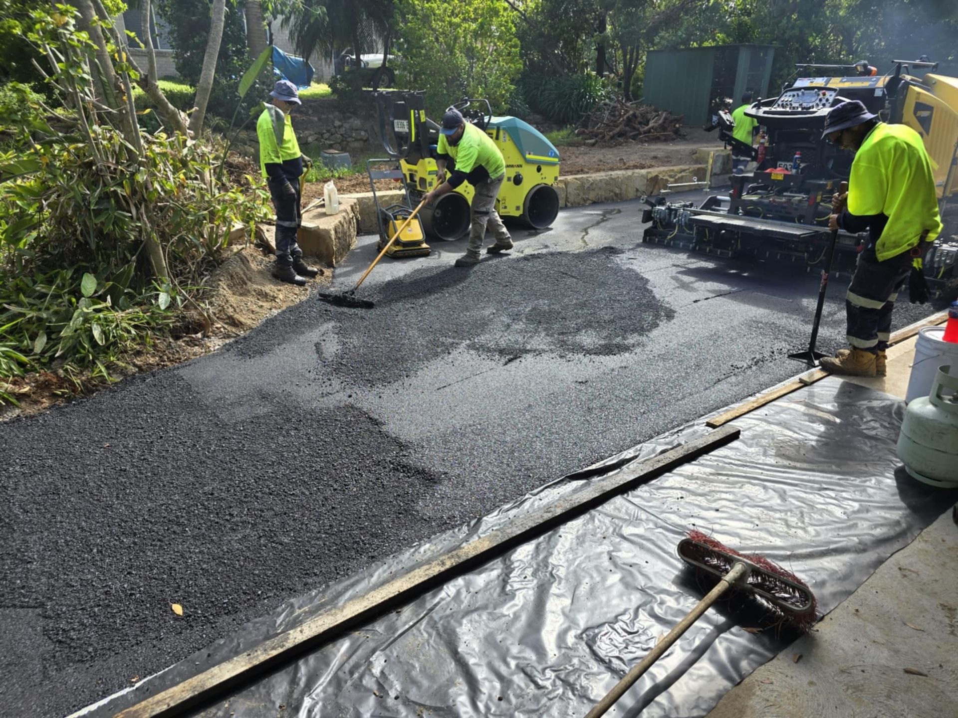 Three construction workers in high-visibility gear paving a driveway with asphalt next to a roller machine.
