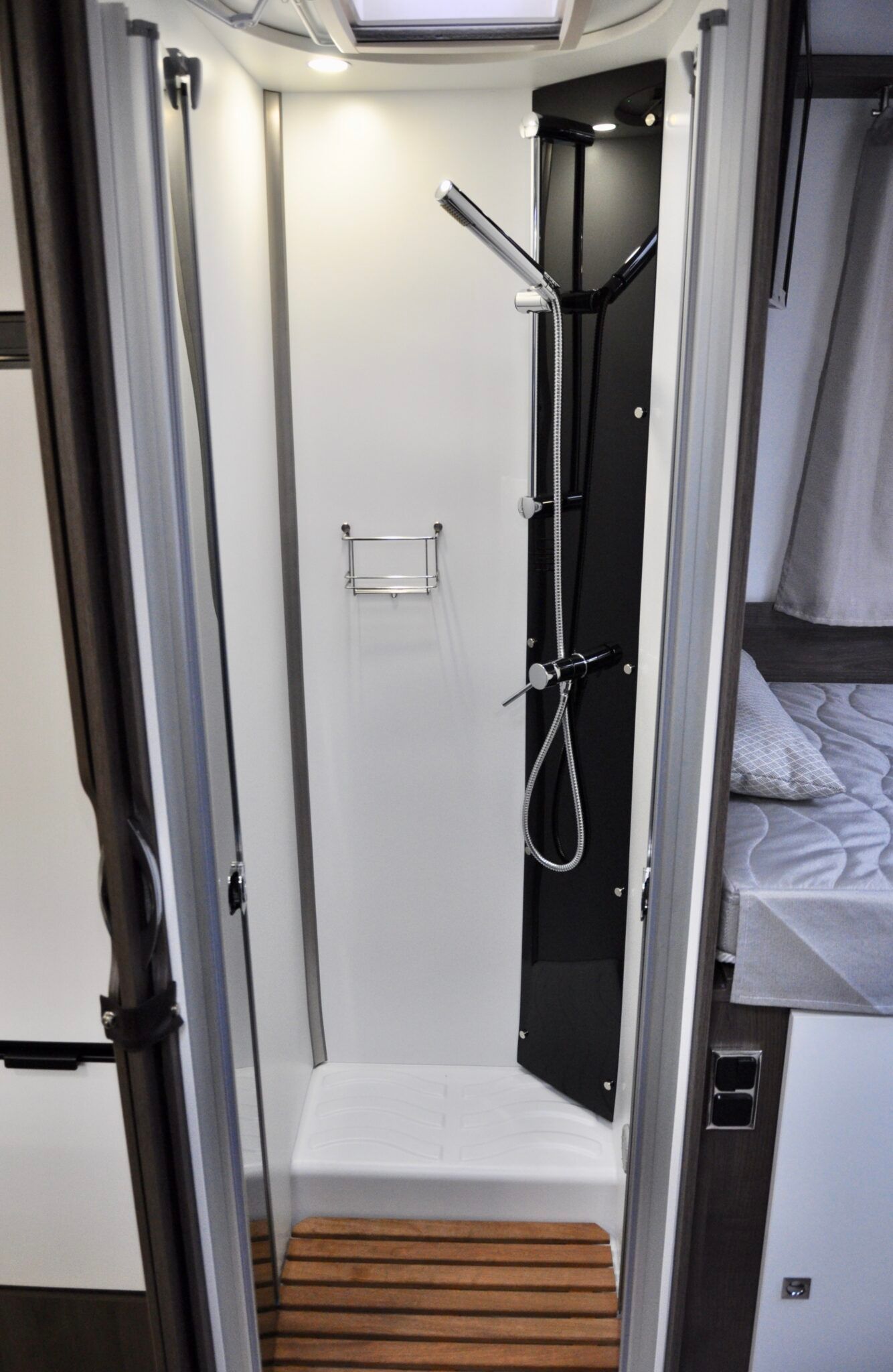 Interieurfoto van een douche in een camper met een houten vloermat en een douchekop op een zwart paneel.