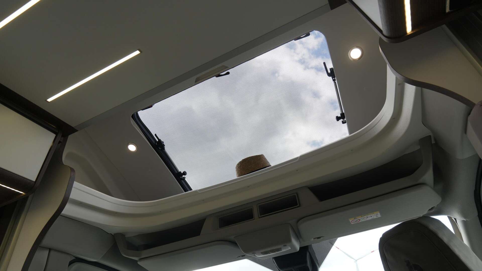 Het interieur van een camperbusje met een open dakraam dat uitzicht biedt op een bewolkte hemel, een opbergplank en verlichting.