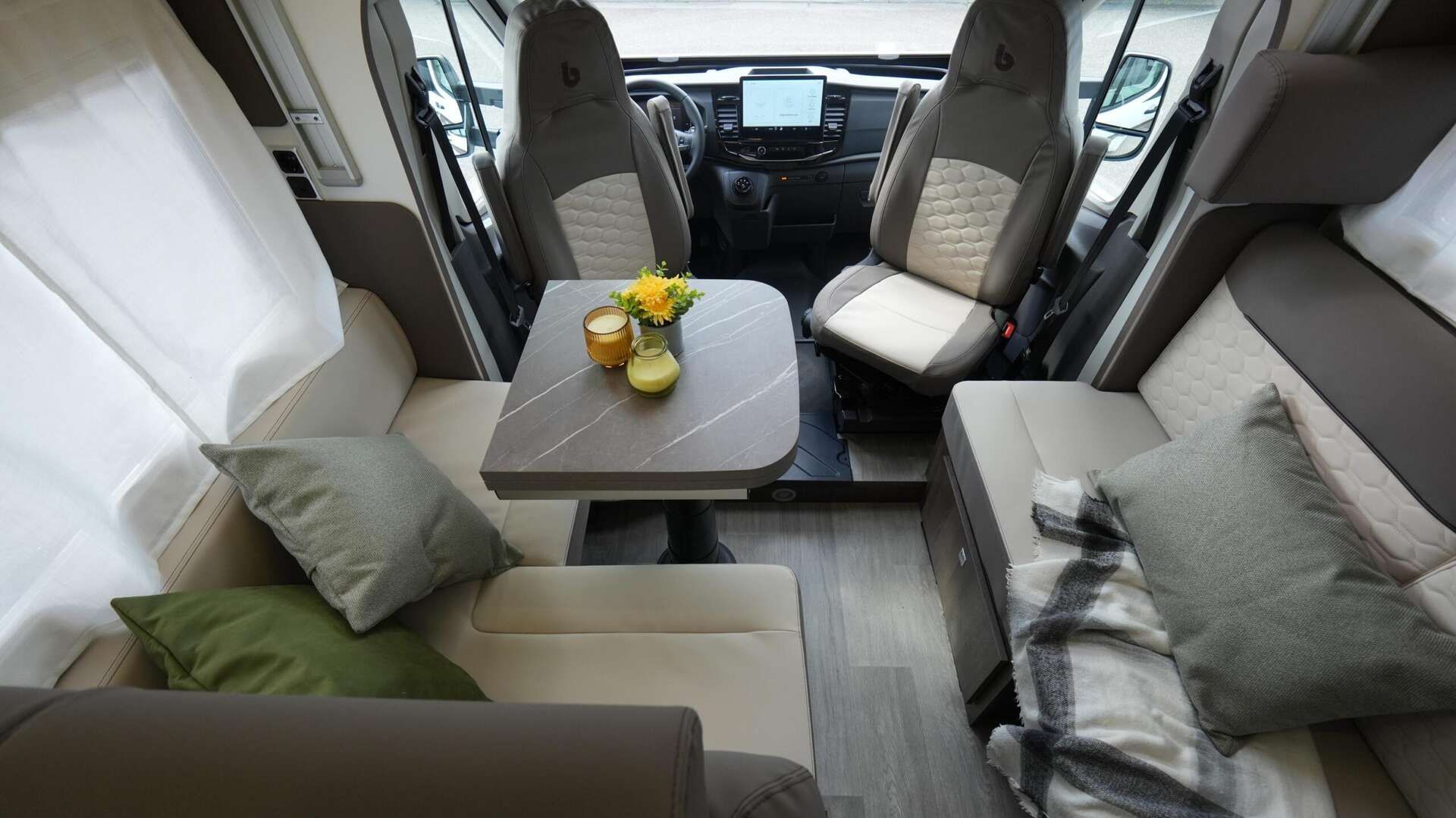 Interieur van een camper met een tafel, zitplaatsen en bestuurdersgedeelte zichtbaar. Beige en grijs kleurenschema.