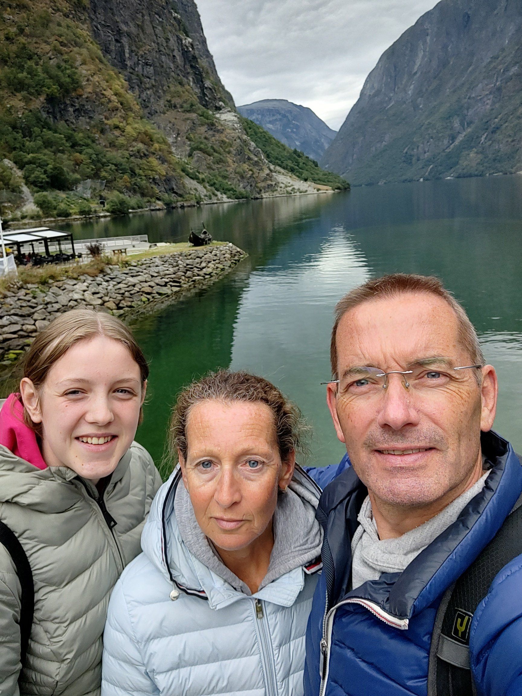 Familieselfie met een schilderachtig fjord op de achtergrond. Bergen, water, bewolkte lucht, groene jassen.