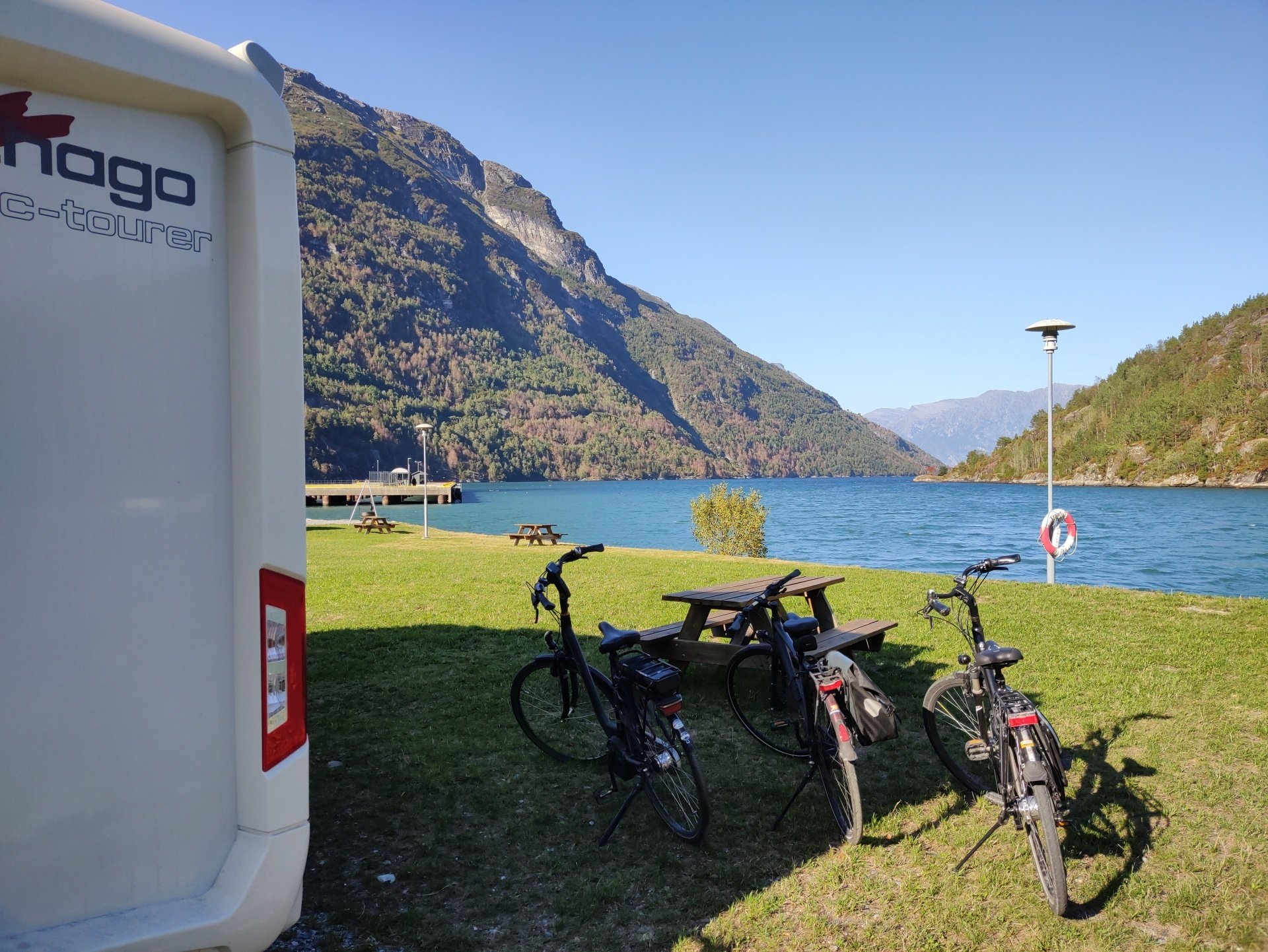 Drie fietsen staan ​​geparkeerd op een grasveldje aan de oever van een meer, met een camper en bergen op de achtergrond.
