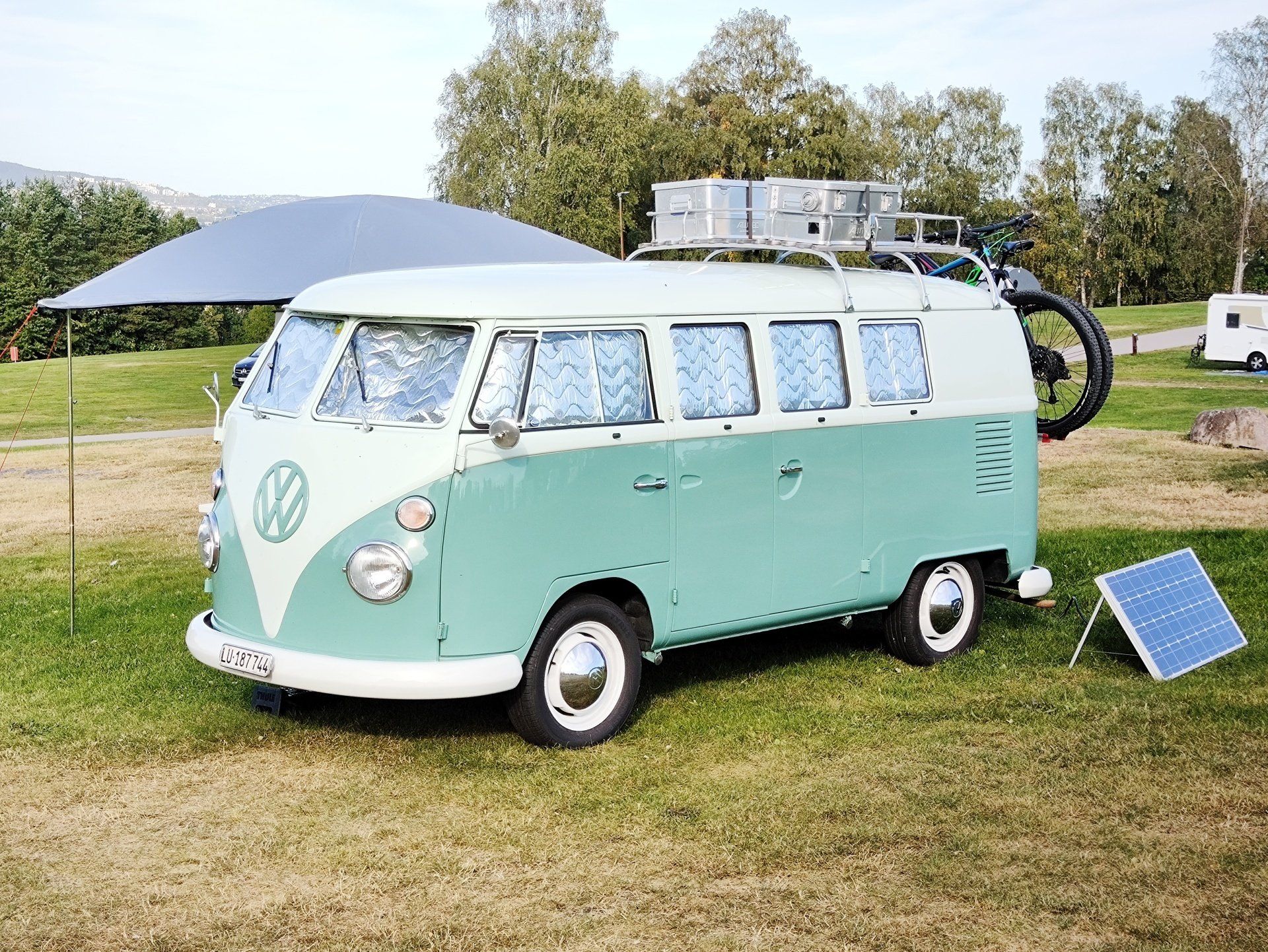 Tweekleurige, turquoise en witte VW-bus met zonnepaneel en fietsendrager, geparkeerd op het gras.