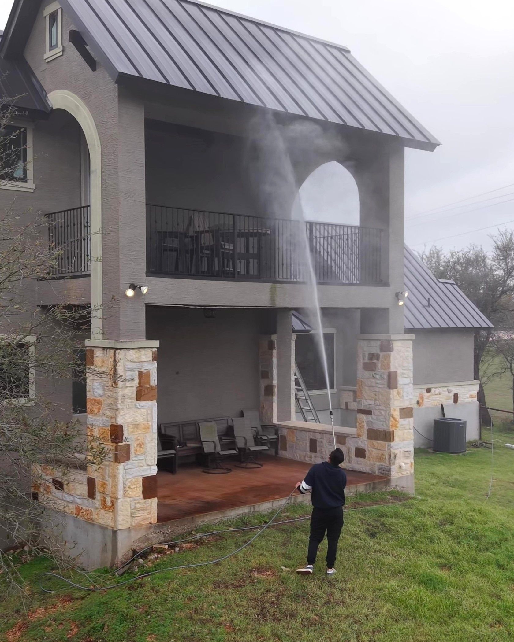 Person pressure washing a two-story house with a balcony, standing in a grassy yard.