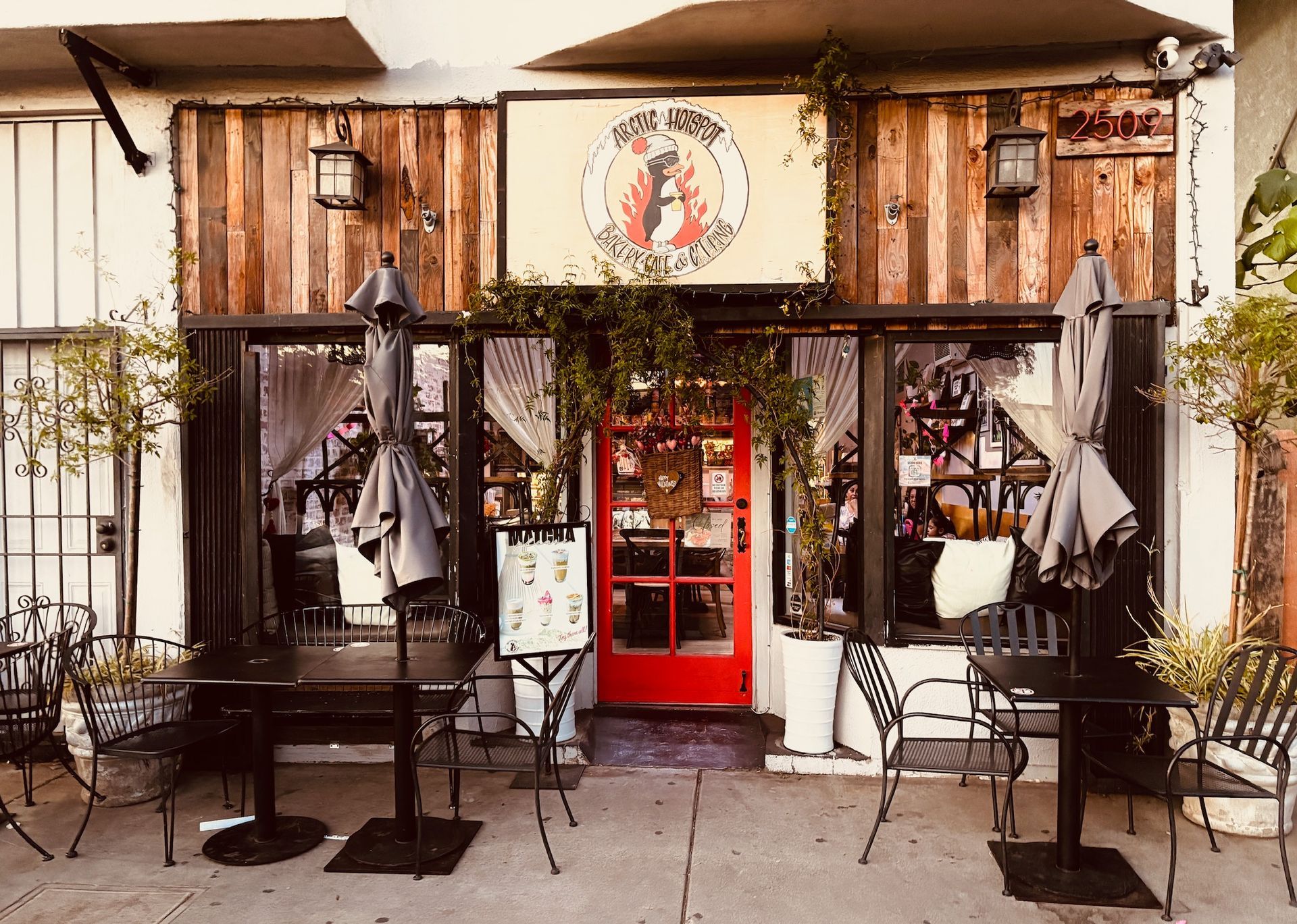 Restaurant exterior with red door, wooden siding, tables, and umbrellas. Sign reads 
