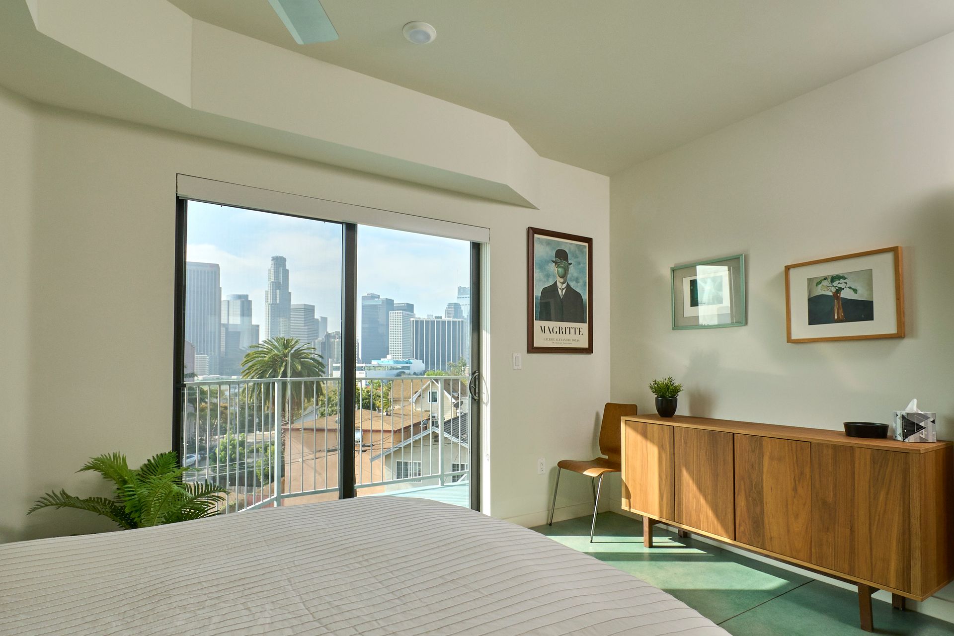 Bright bedroom with balcony view of city skyline, green bedspread, and wooden dresser with framed art