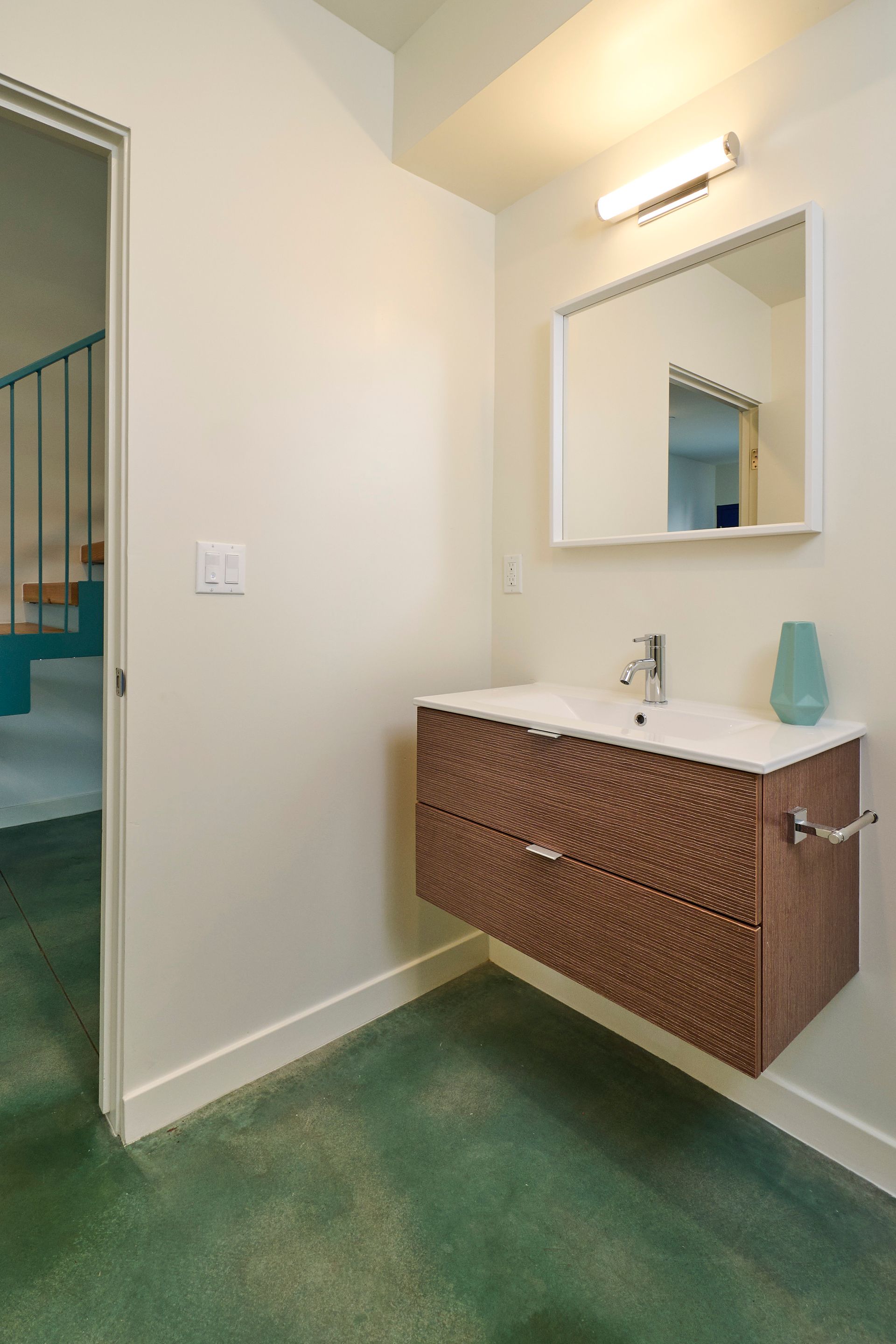 Small bathroom vanity with mirror, white walls, woven wood cabinet, and green carpeted floor.