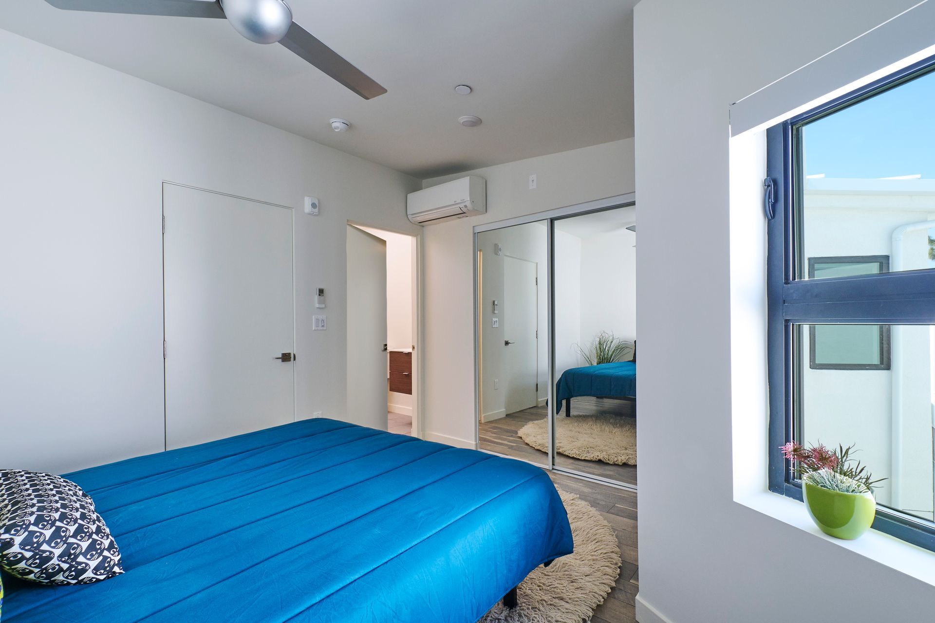 Bright bedroom with blue bedspread, mirrored closet, and window overlooking a city view.