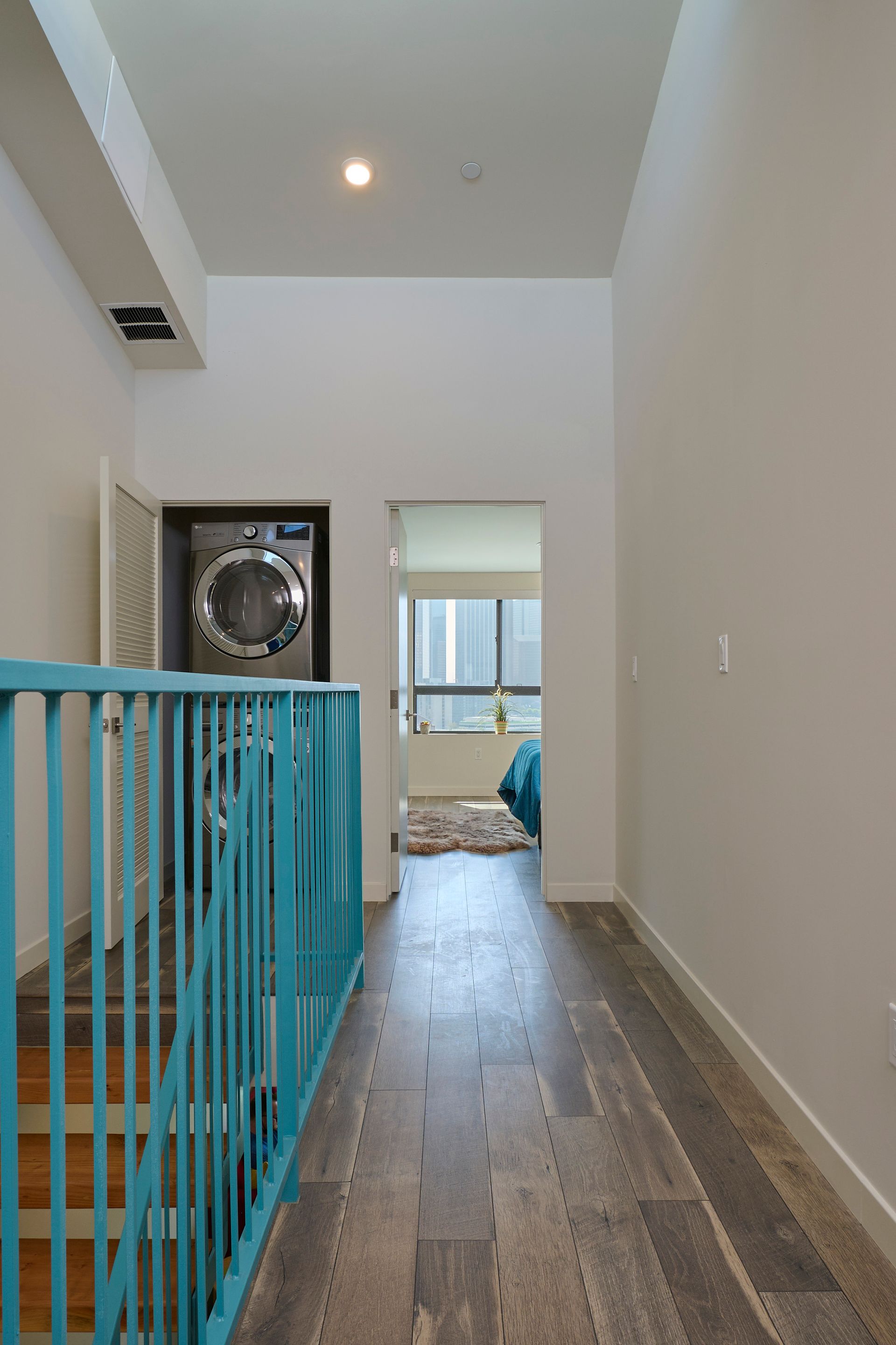 Narrow hallway with wood flooring, blue railing, and laundry room visible at the end.
