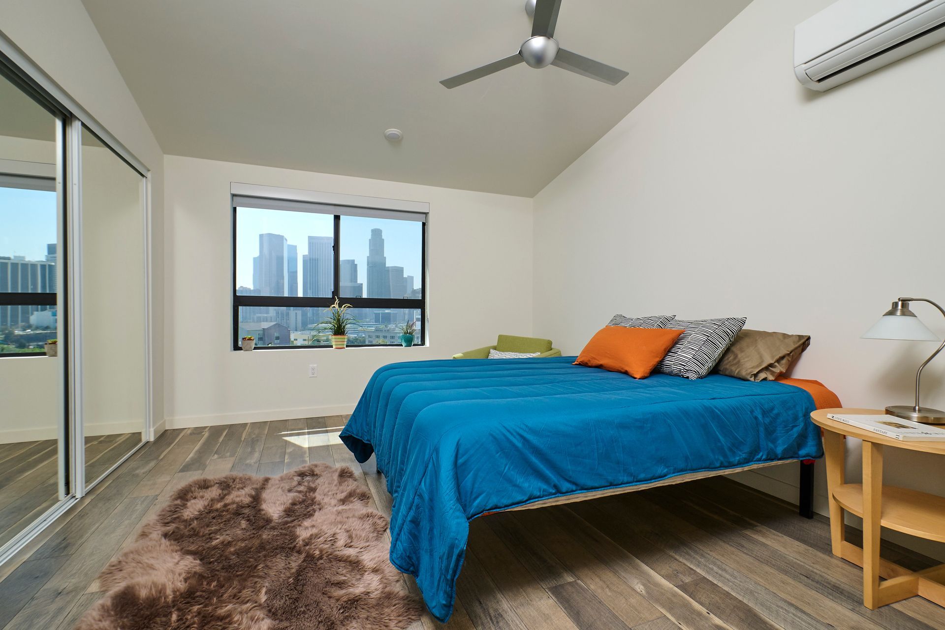 Bright bedroom with blue bedspread, window city view, mirrored closet, and ceiling fan