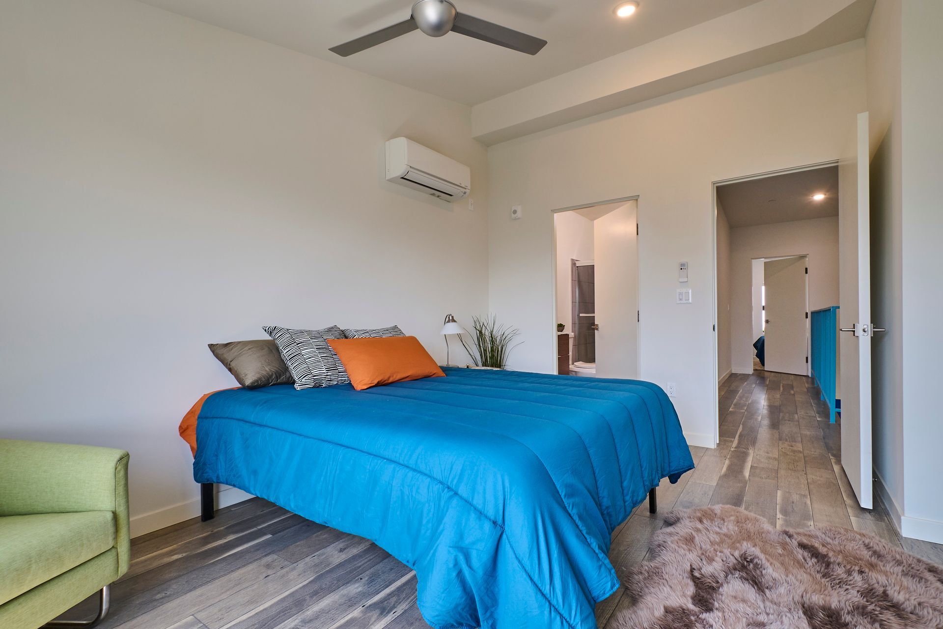 Bedroom with a blue bedspread, green chair, ceiling fan, and hallway view.