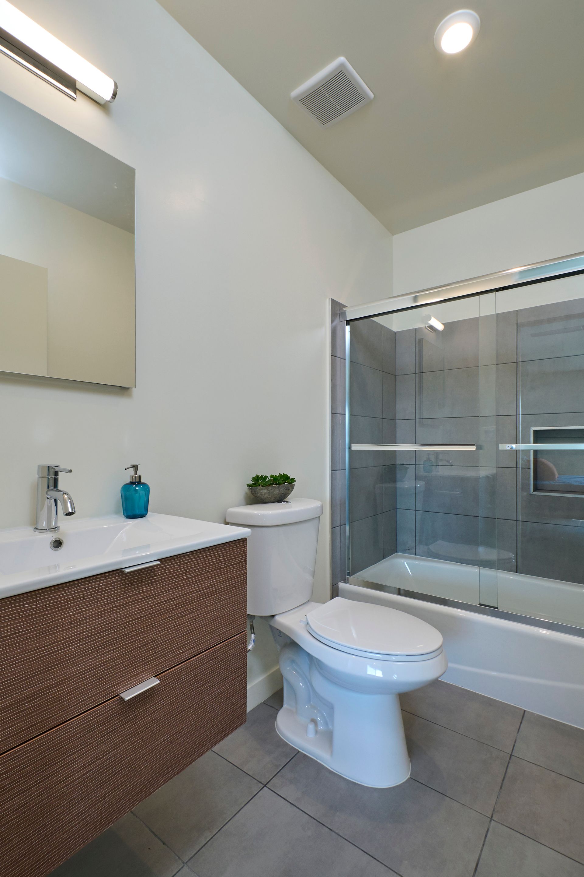 Modern bathroom with white toilet, glass shower, wicker vanity, and gray tile floor.