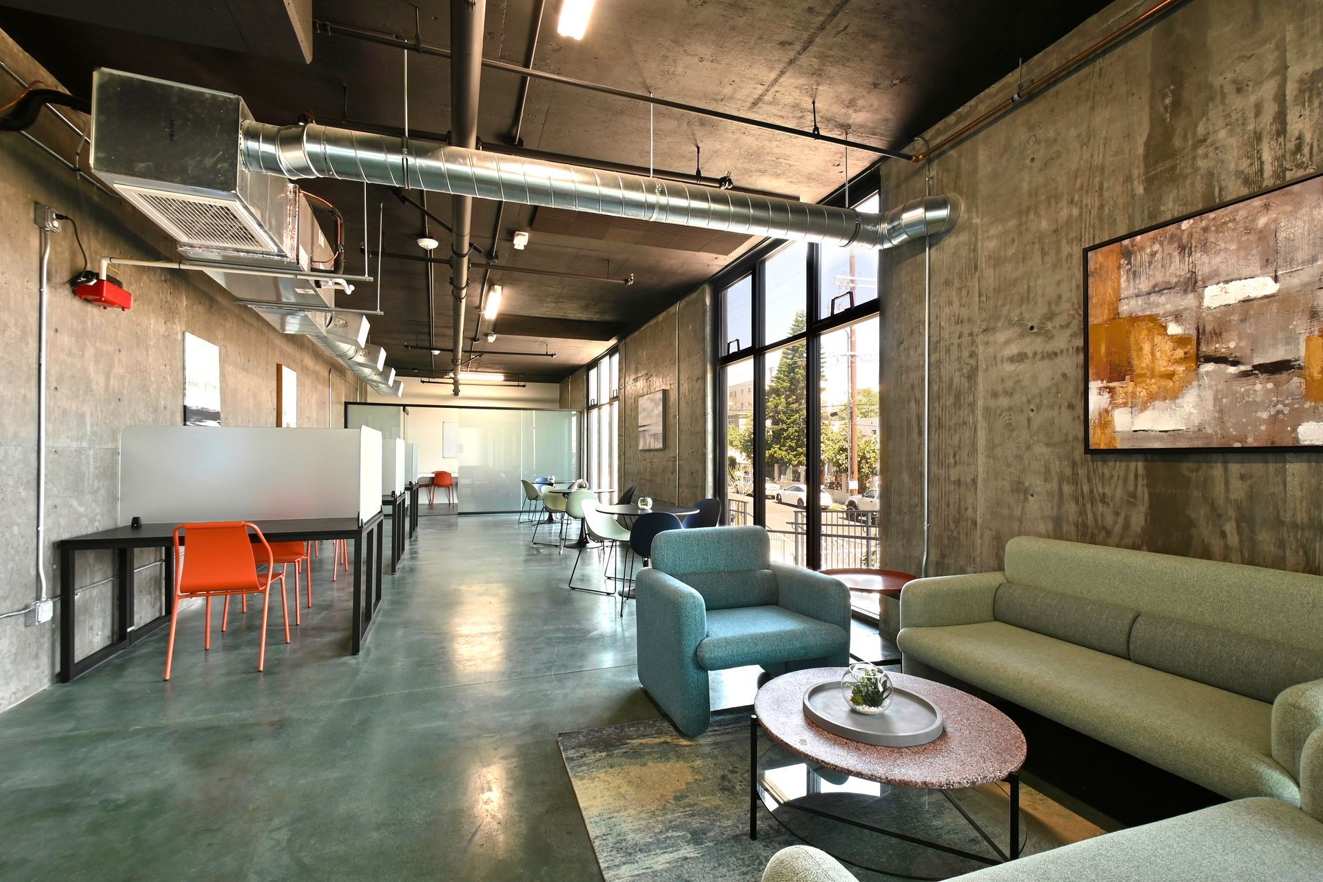 Modern loft-style lounge with green sofas, orange chairs, exposed ceiling, and large windows.