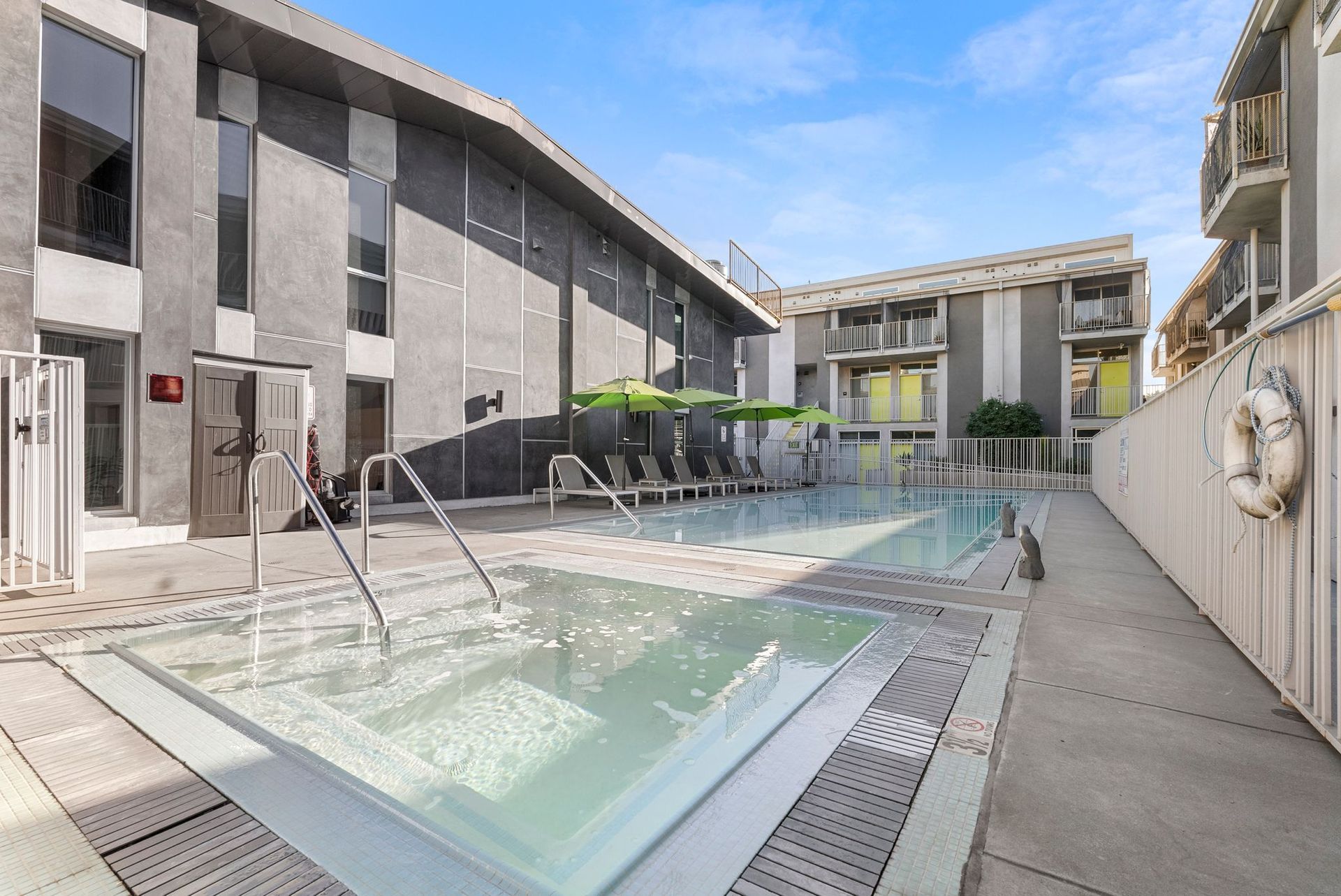 Swimming pool area with pool, spa, lounge chairs, and buildings.