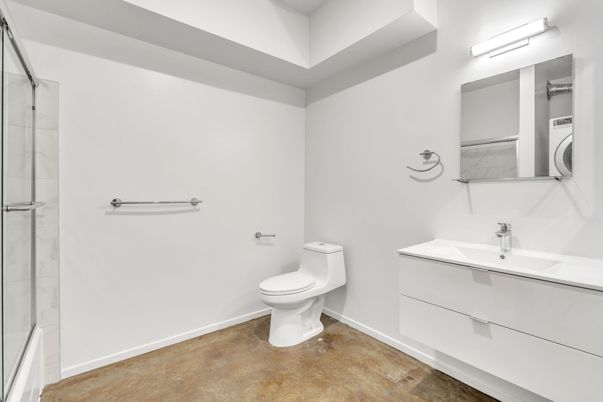 White bathroom with a toilet, sink, mirror, and shower. Concrete floor and towel racks.