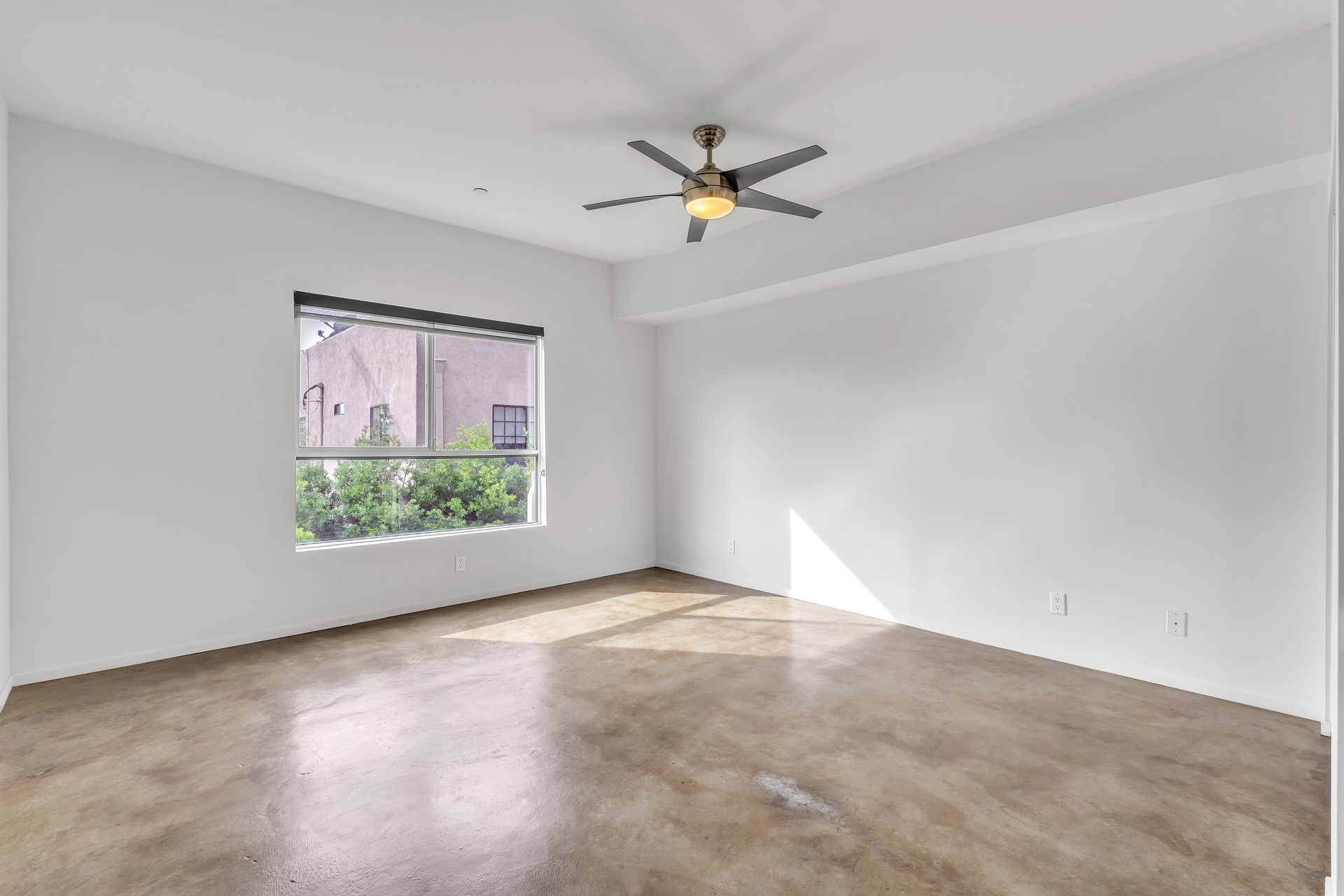 Empty room with a window, ceiling fan, and concrete floor. Bright light, white walls.