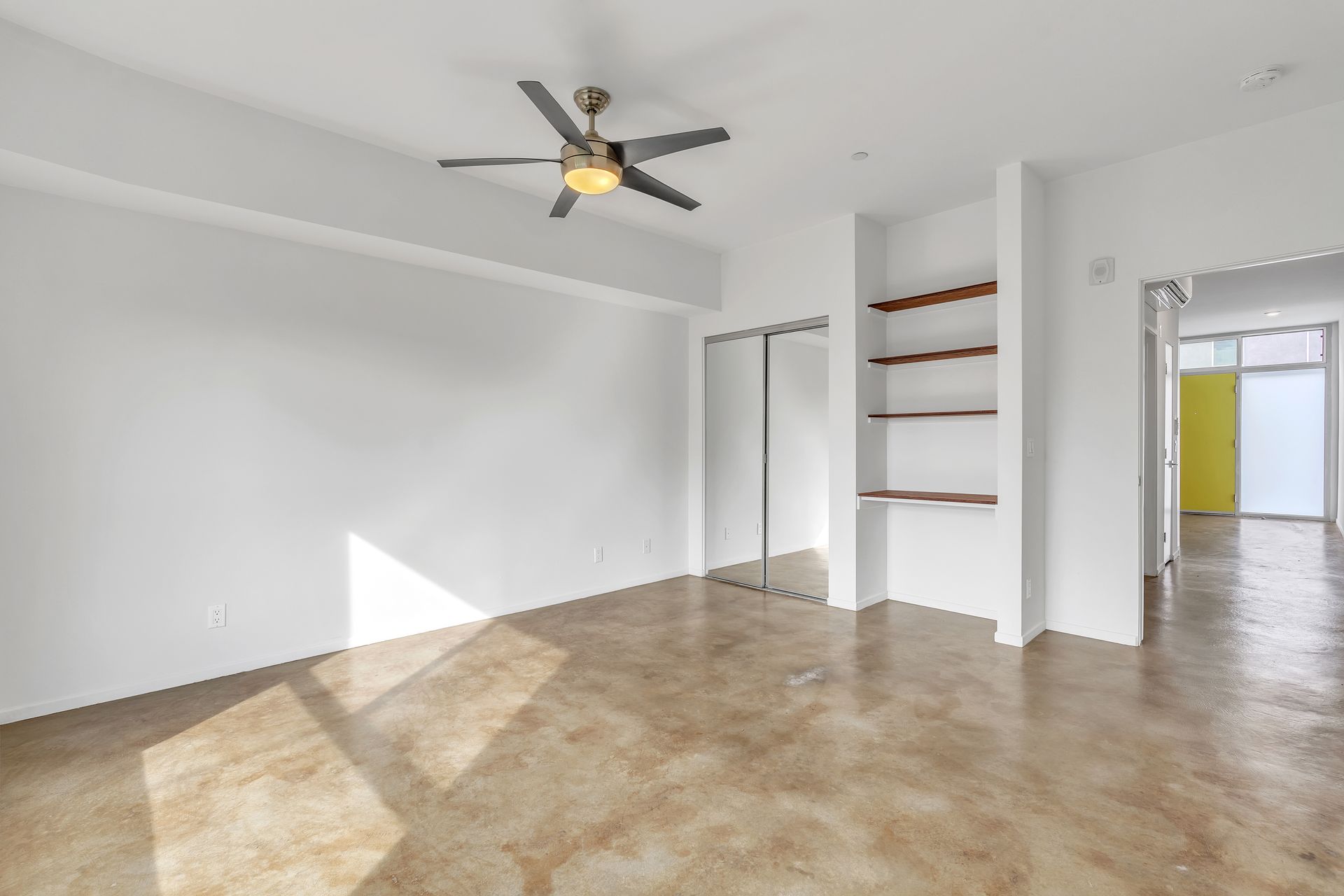 Empty room with a ceiling fan, built-in shelves, and a sliding door. Sunlight streams in.