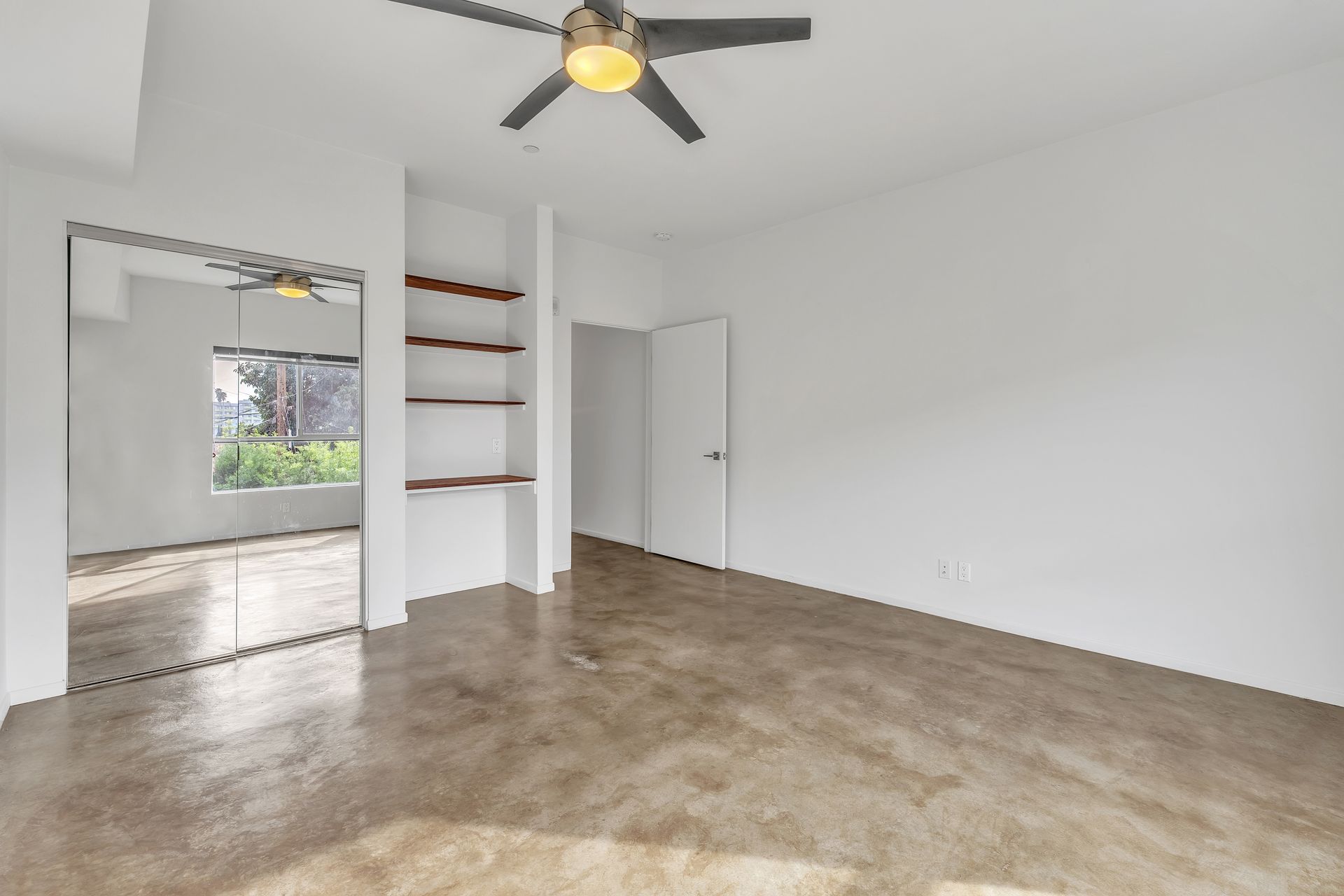 Empty room with polished concrete floor, white walls, built-in shelves, doorway to another room, ceiling fan.