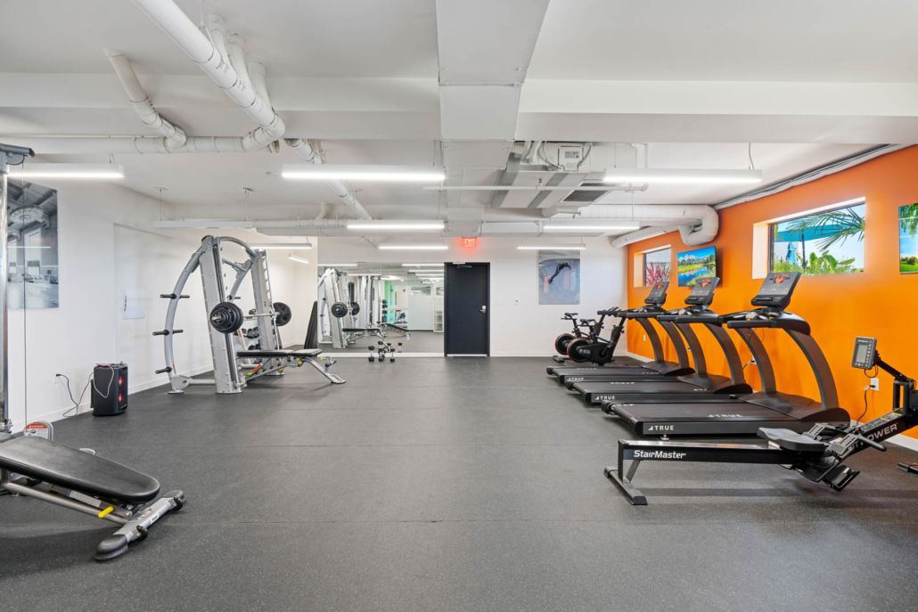 Empty gym with cardio machines, weight equipment, and bright orange accent wall under white ceiling lights