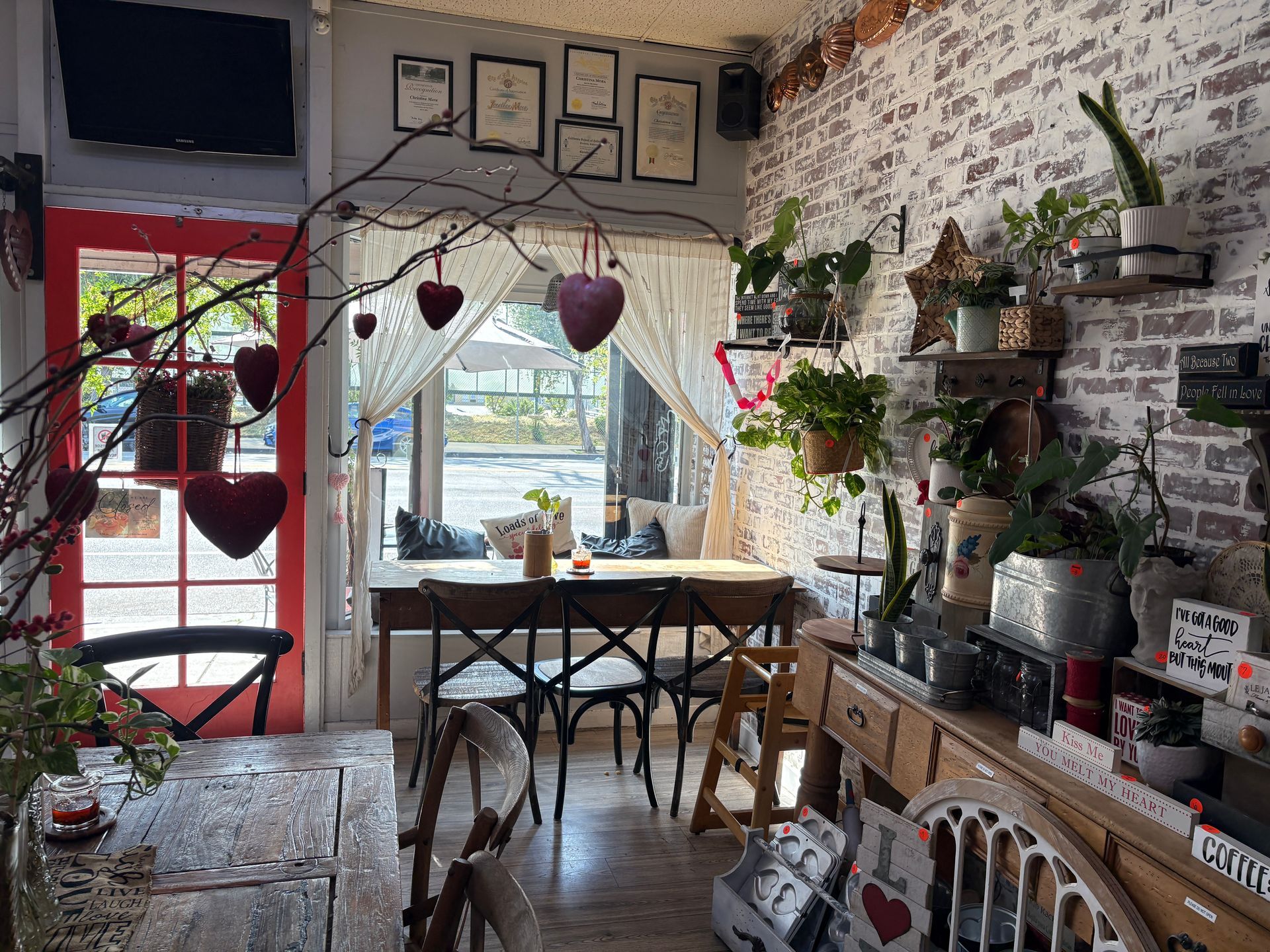 Cozy cafe interior with red door, brick wall, plants, heart decorations, and window table with chairs.