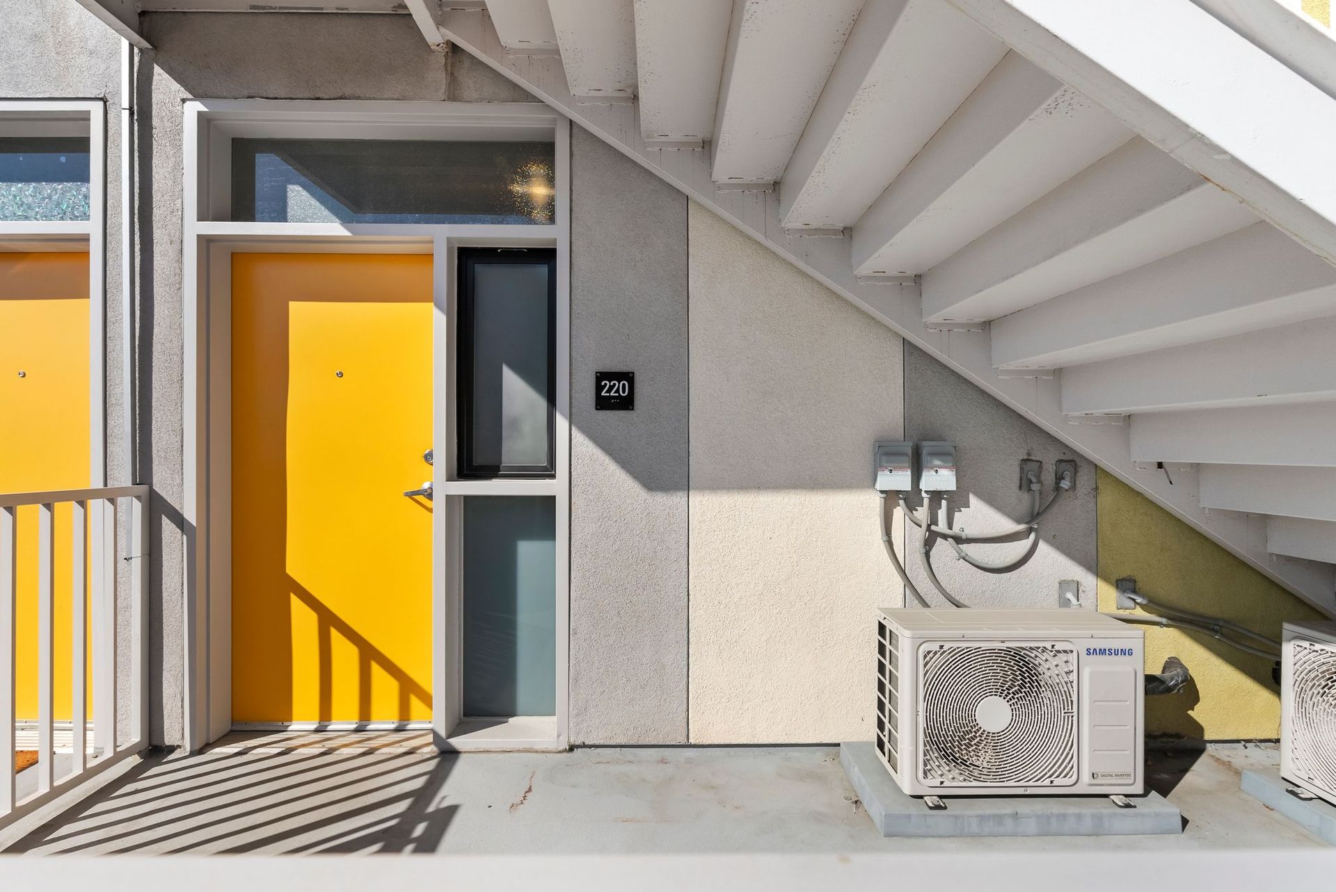 Yellow apartment doors under an exterior staircase, with a utility unit and railing on the right