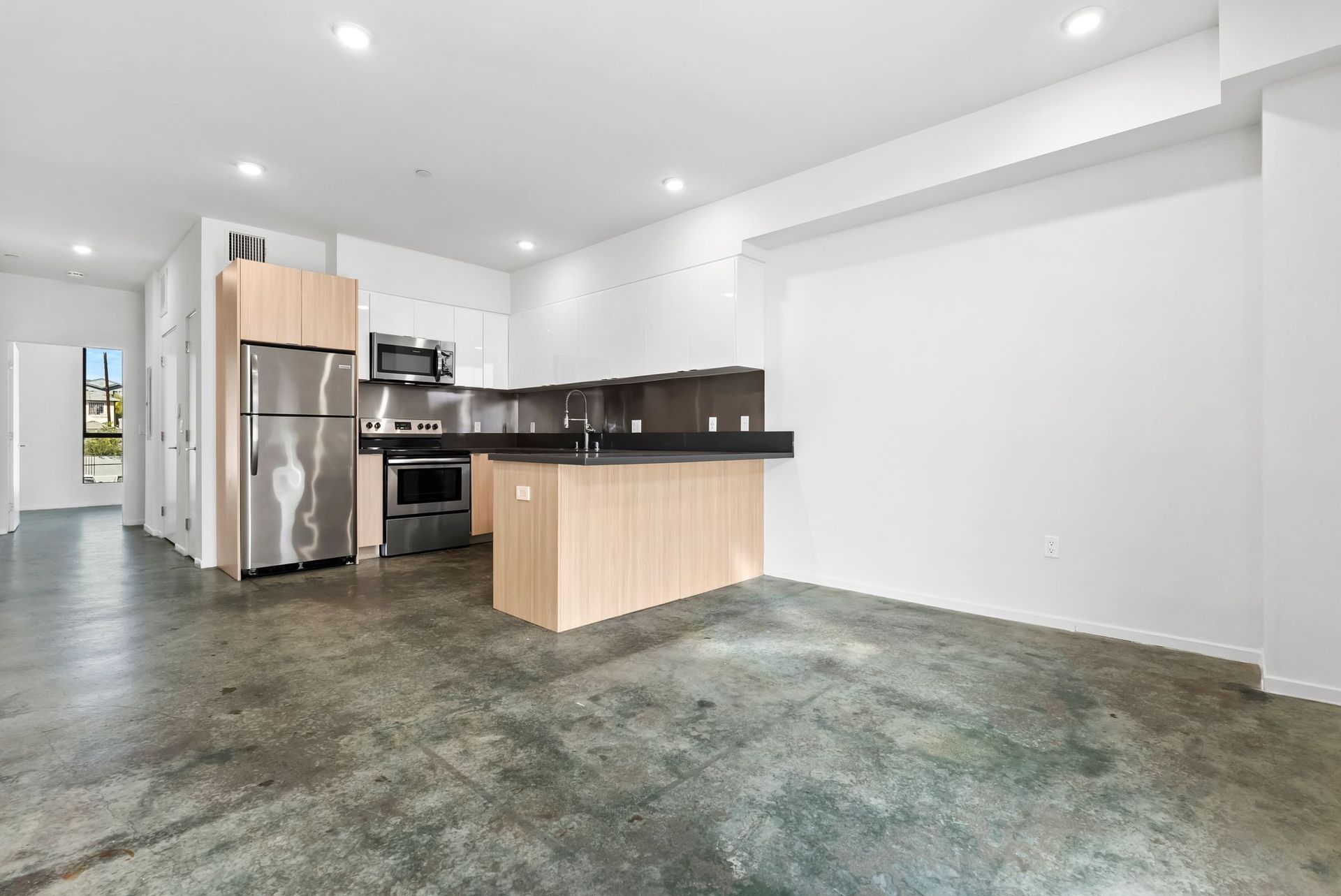 Empty modern kitchen with stainless steel appliances, wood cabinets, and an unfinished concrete floor