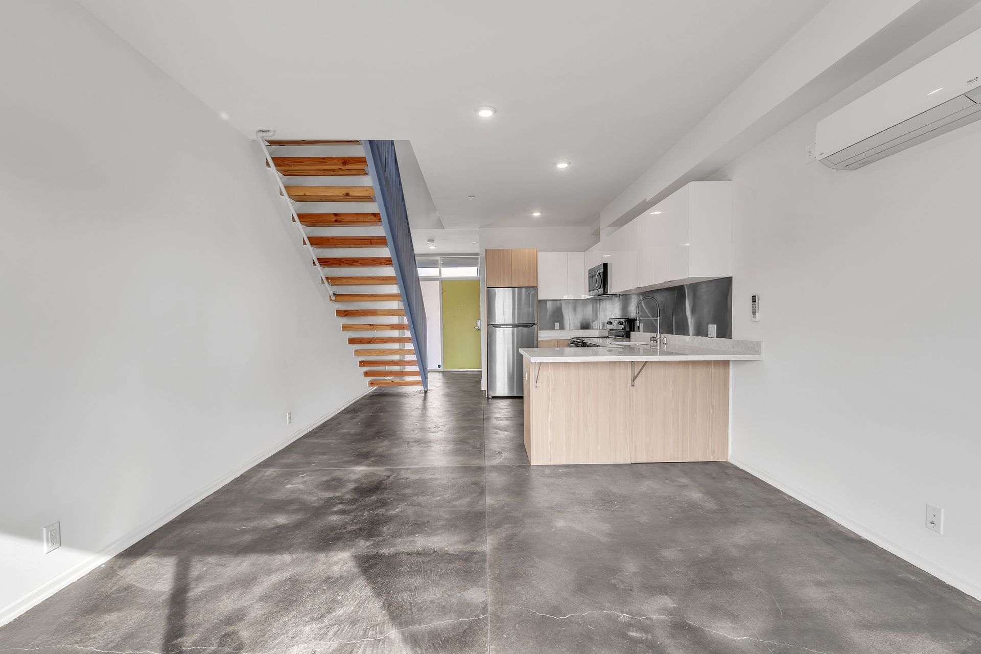 Modern, empty apartment with concrete floor, white walls, kitchen, and wooden staircase.