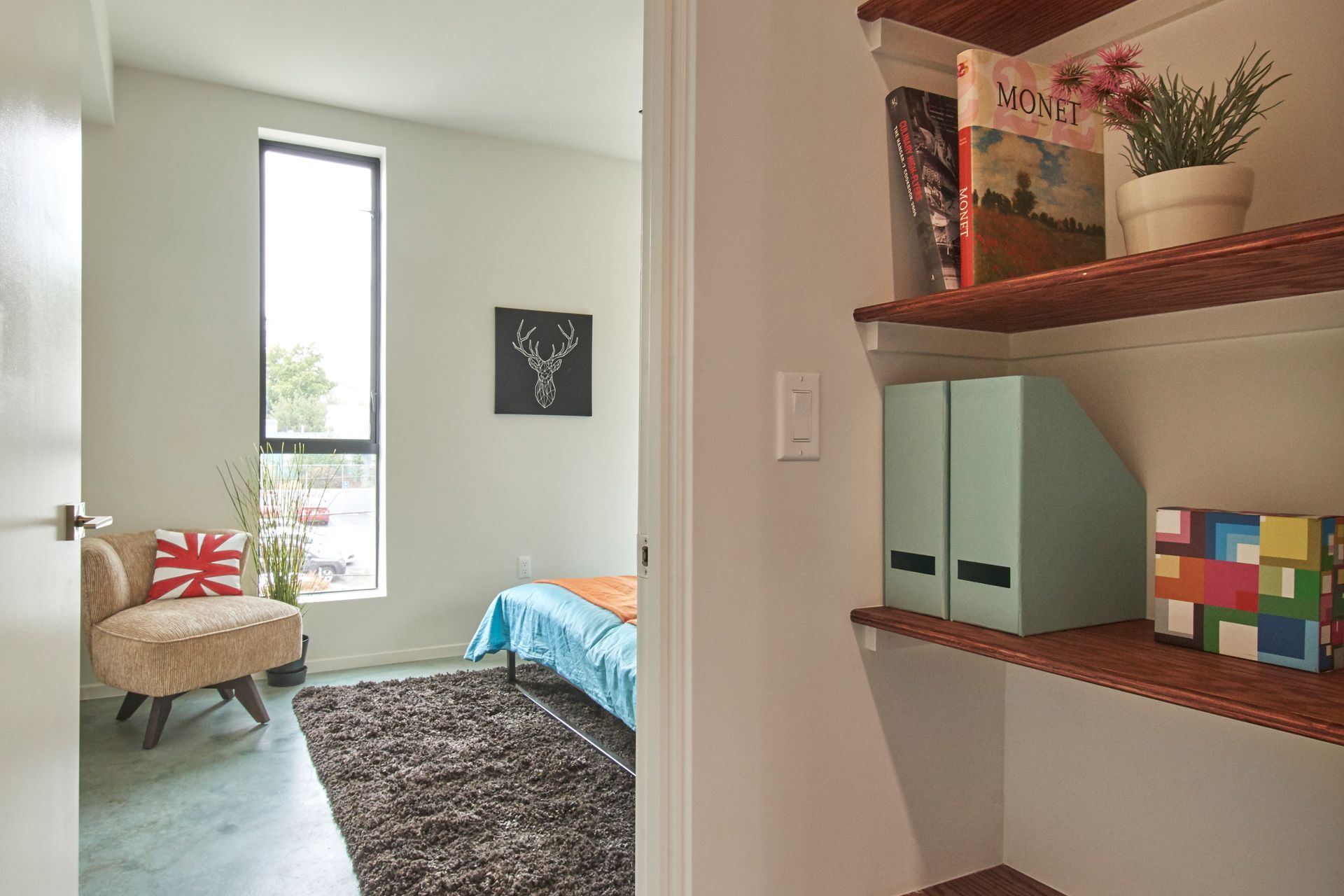 Small bedroom with bed, chair, window, and wall shelves holding decor and storage boxes.