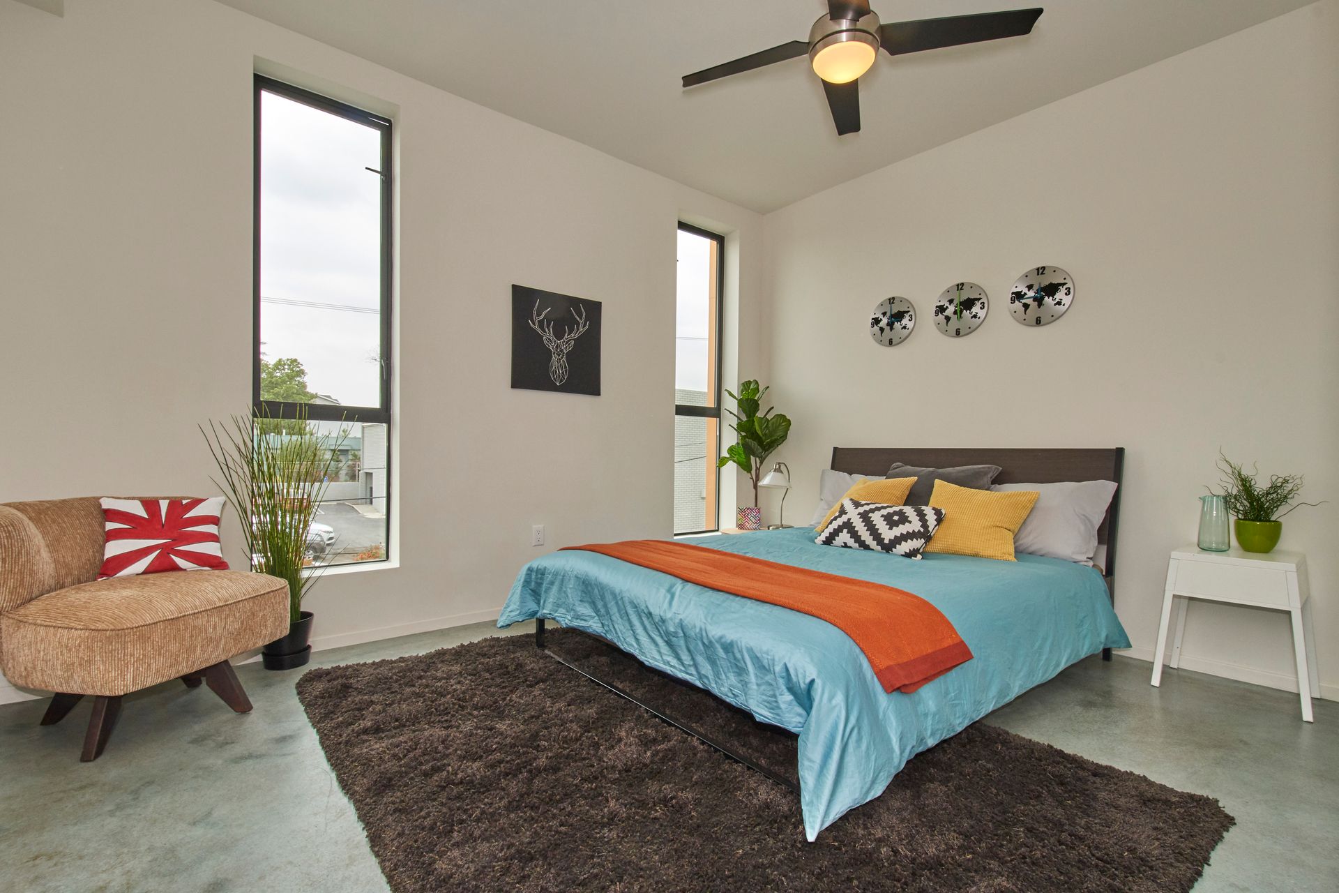 Modern bedroom with turquoise bedspread, dark rug, ceiling fan, and tall windows