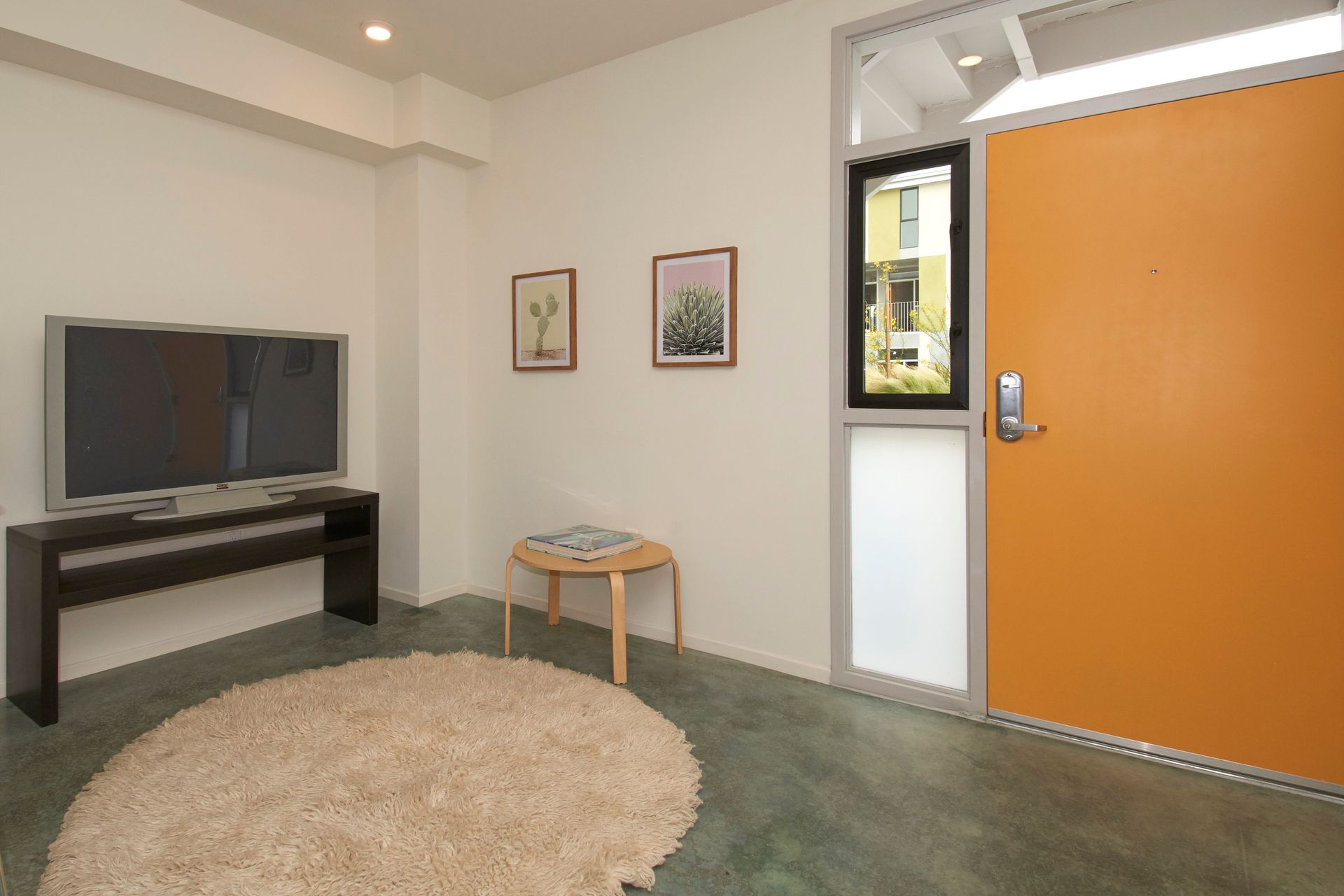 Small living room with TV, rug, table, framed art, and an orange front door open to outside.