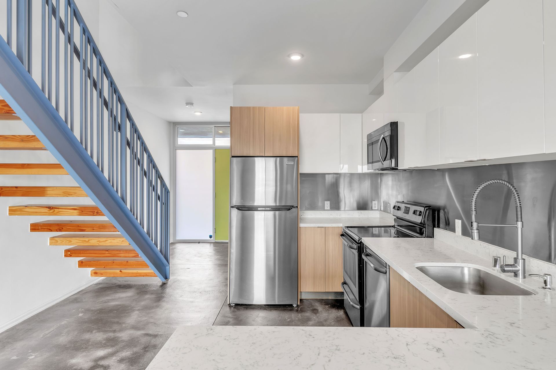 Modern kitchen with stainless steel appliances, light wood cabinets, and a blue staircase.