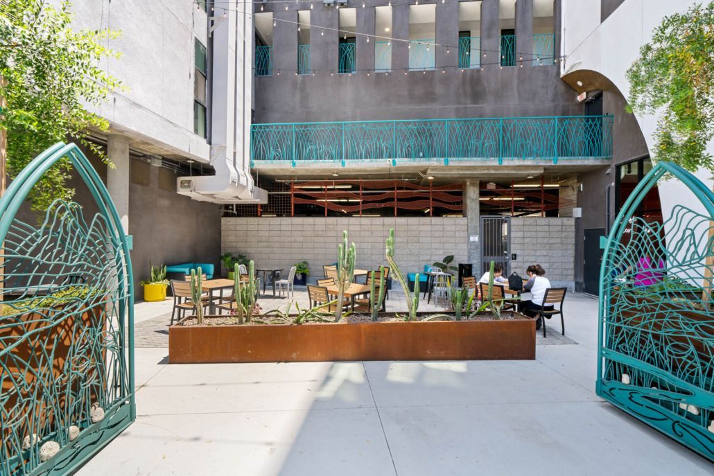 Outdoor courtyard dining area with tables and chairs, framed by turquoise decorative arches and a modern building.