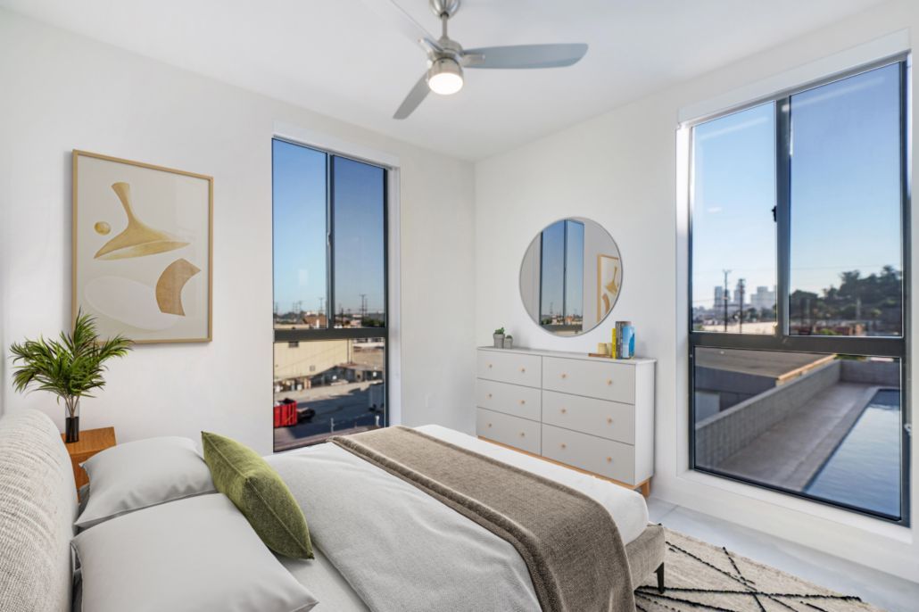 Bright bedroom with bed, dresser, round mirror, and two large windows overlooking a city view