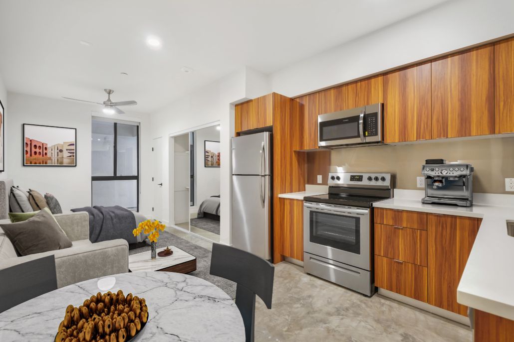 Bright modern kitchen and living room with stainless appliances, wood cabinets, gray sofa, and round table