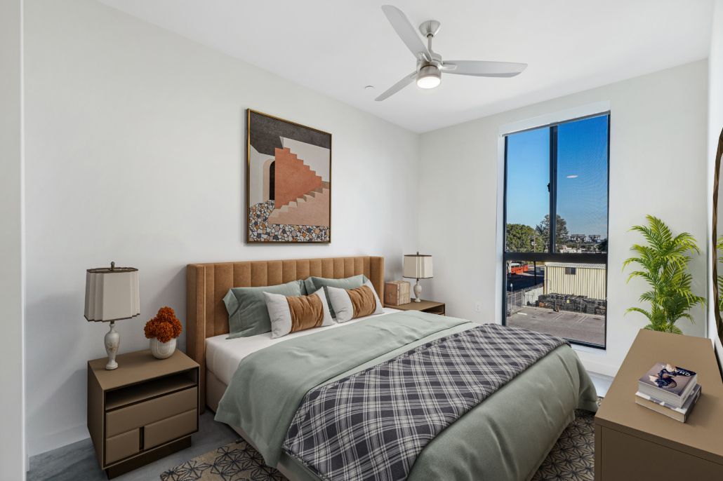 Bright bedroom with a bed, nightstands, ceiling fan, and a window overlooking a city view.