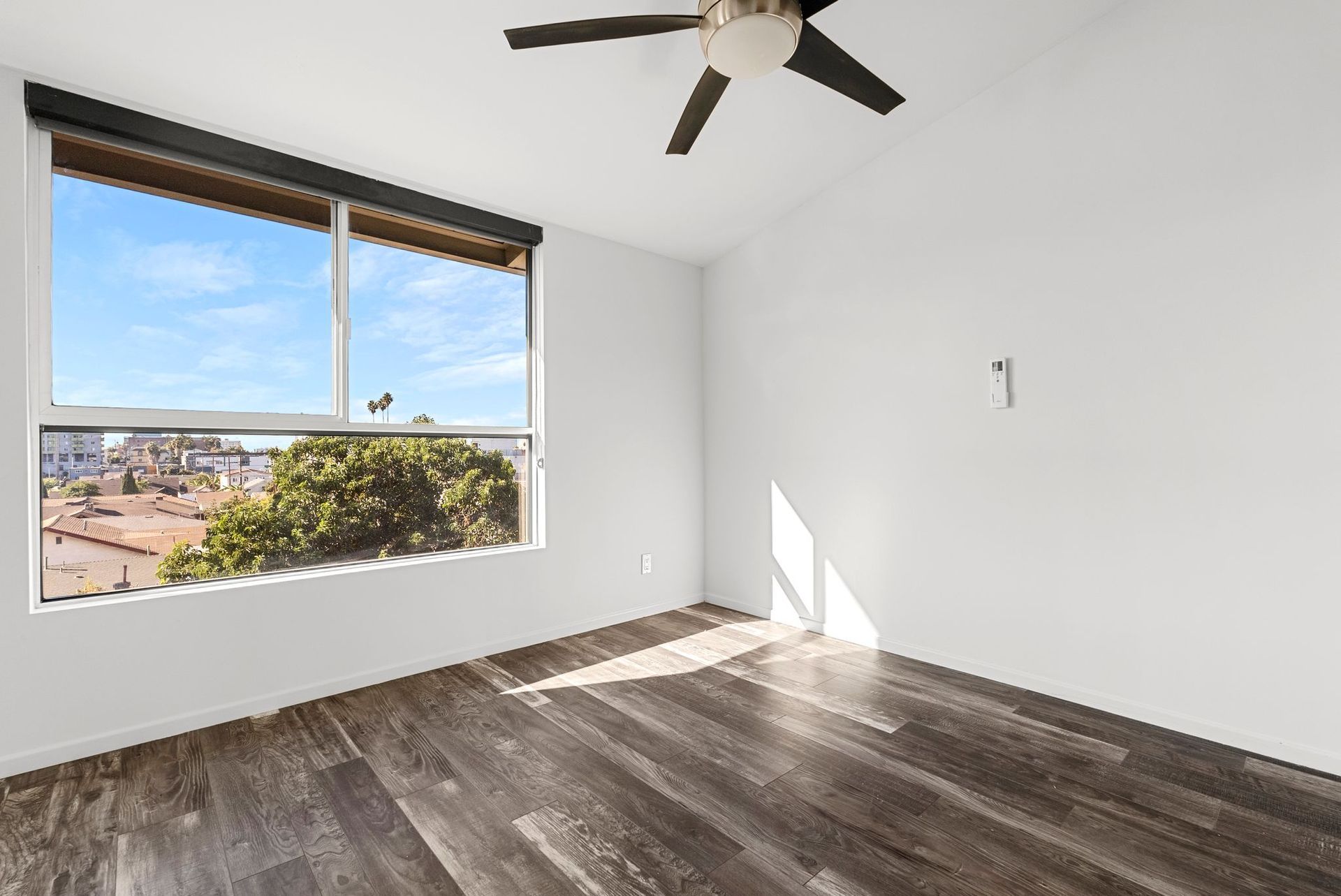 Empty room with dark wood floors, large window, white walls, and a ceiling fan.