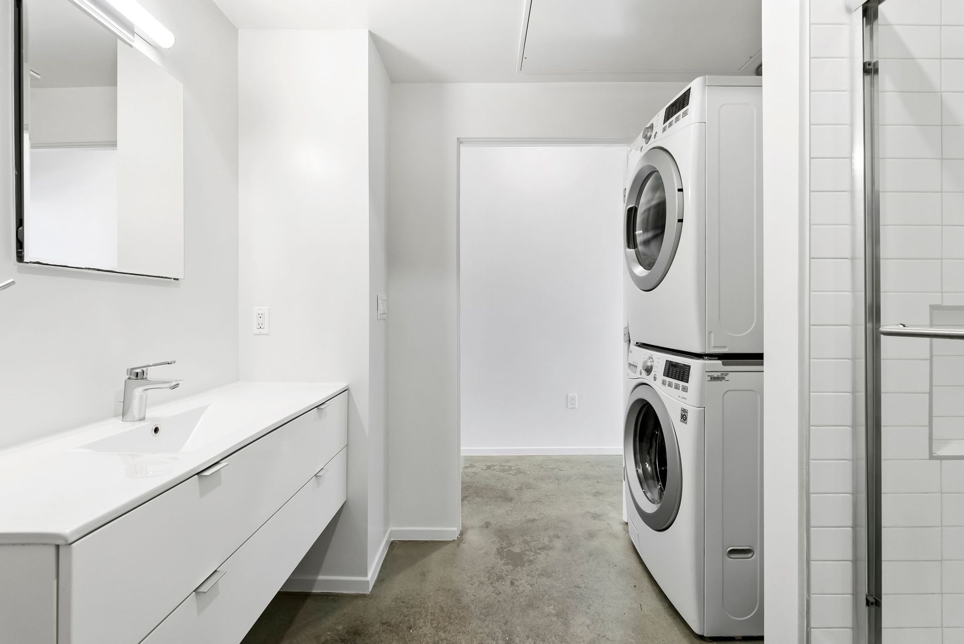Modern white laundry room with stacked washer and dryer, sink, and glass shower door