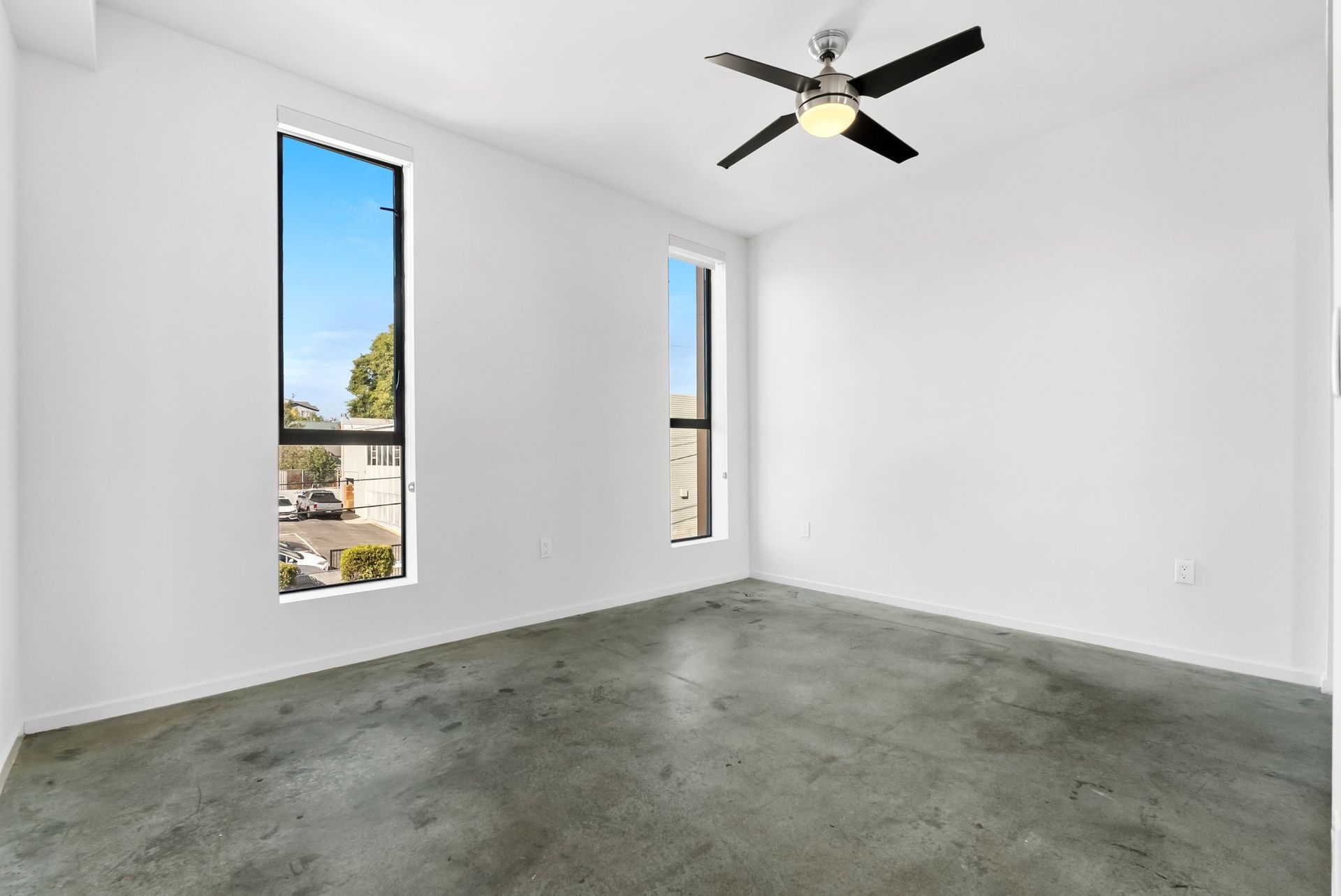 Empty white room with gray floor, two tall windows, and a ceiling fan