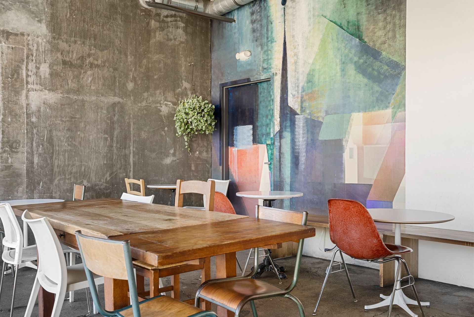 Cafe interior with wooden tables and mismatched chairs against a concrete wall and colorful mural.