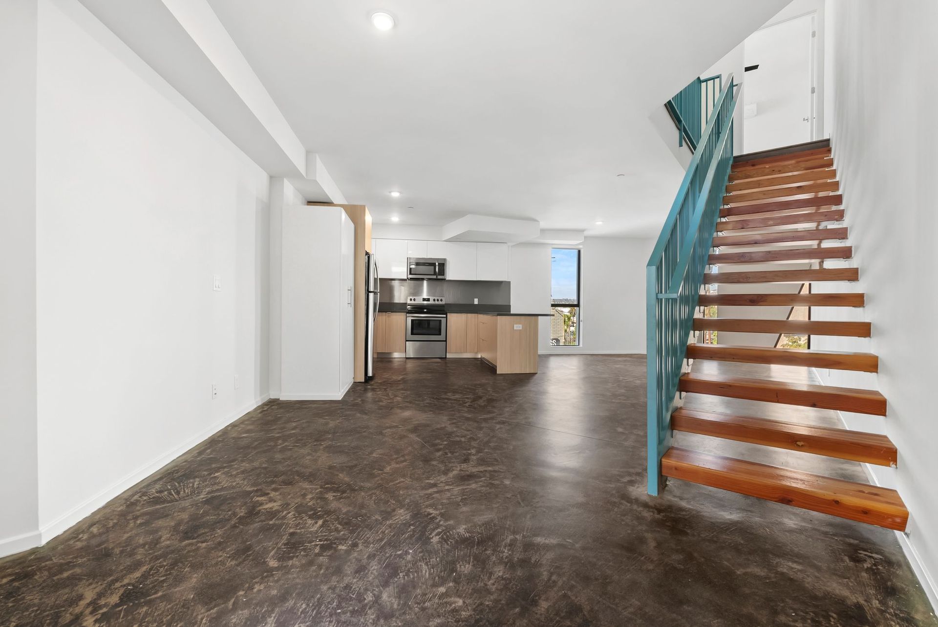 Modern empty open-plan interior with kitchen, dark floors, and a staircase with teal railing