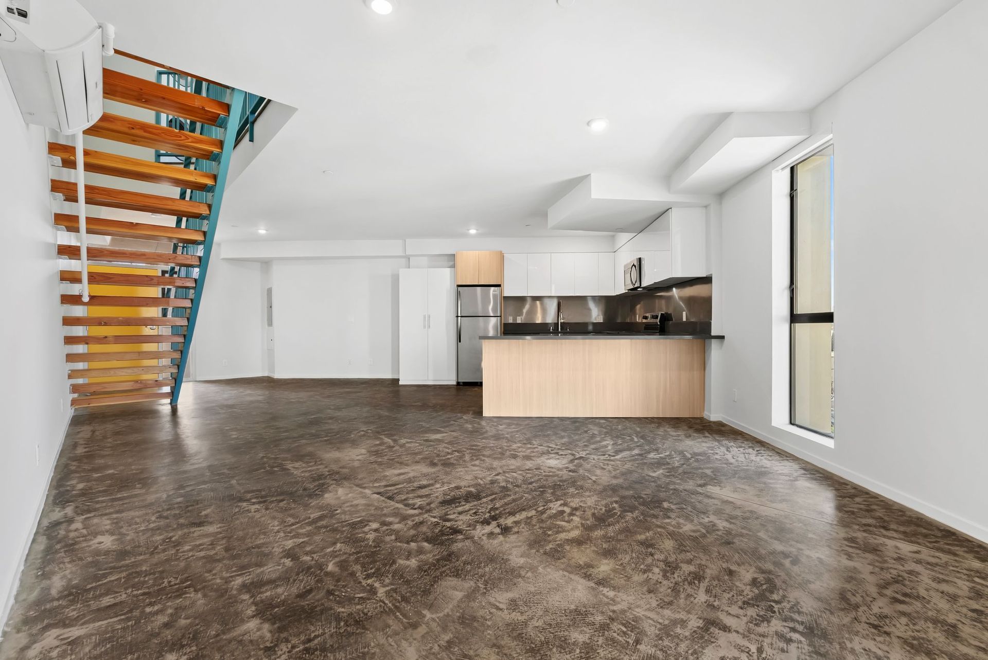 Empty modern loft with wood staircase, open kitchen, white walls, and polished concrete floor